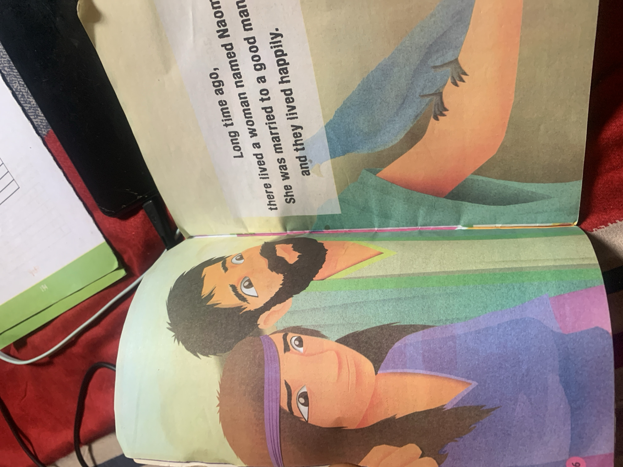 The image shows an open book with illustrations and text. The left page features an illustration of a man and a woman. The man has dark hair and a beard, and is wearing a green tunic. The woman has dark hair with a purple headband and is wearing a purple top. The right page displays text that reads: "Long time ago, there lived a woman named Naomi. She was married to a good man and they lived happily." Below the text, there is an illustration of a blue bird perched on an arm. The background of the book's pages is a gradient of green. The book is placed on a red surface, possibly fabric, with some wires and a green folder visible in the upper portion of the image. The image does not provide any information about the location in the city of Jauro Yinu, Nigeria.