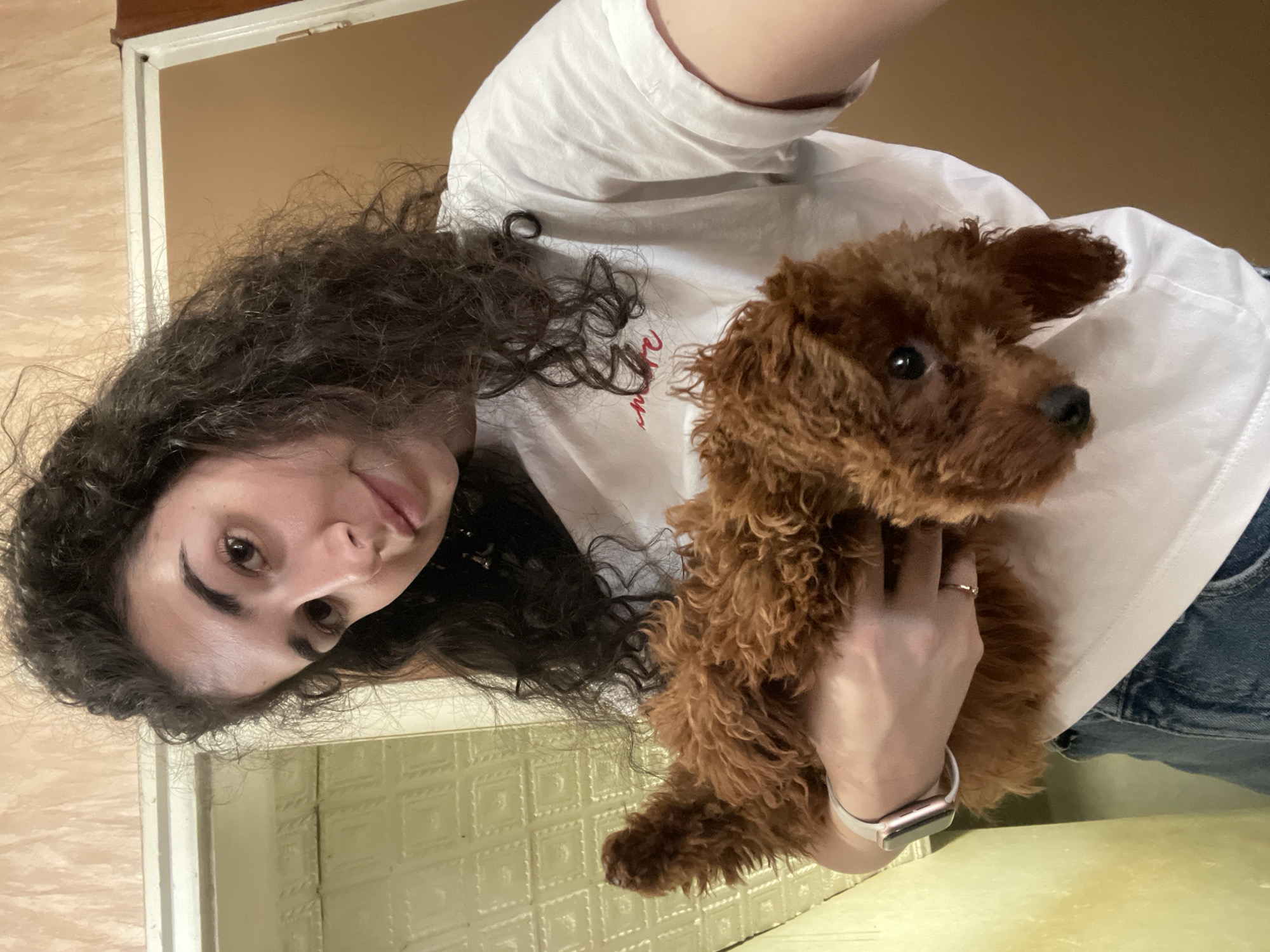 A woman with curly, dark brown hair and fair skin is holding a brown poodle in her arms. She is wearing a white t-shirt and blue jeans. She is wearing a ring on her finger and an Apple watch. She appears to be inside a building, near a beige-colored wall with white trim. The location is unknown, but the city is specified as Sofia, Bulgaria.