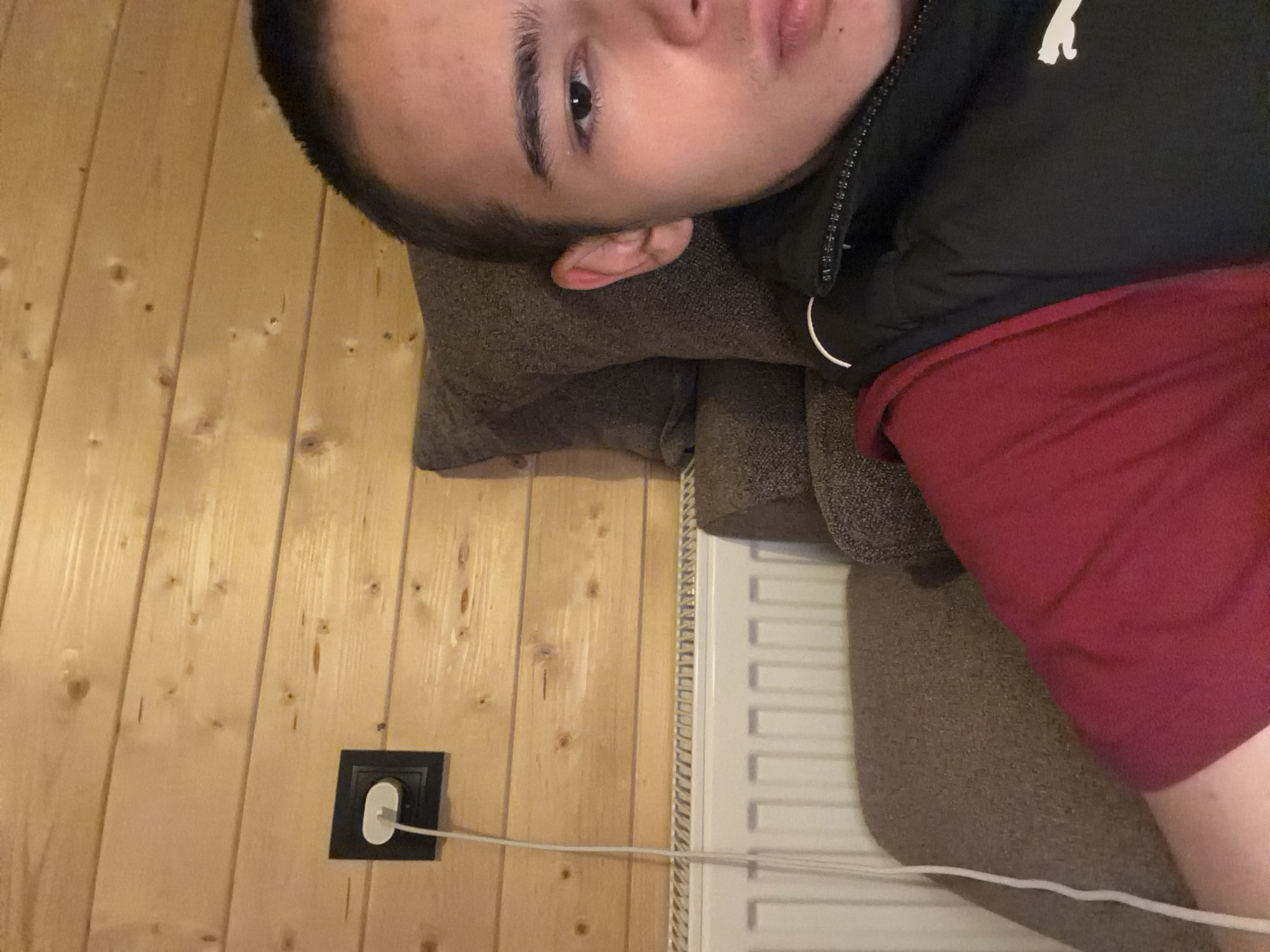 An indoor, medium shot photograph shows a young person from the chest up, reclining on a brown sofa with a grey pillow behind their head. The person has short dark hair and is wearing a maroon t-shirt and a black vest. The background consists of a wood-paneled wall with a black electrical outlet and a white cord plugged into it. A white radiator is visible below the wall. The lighting is dim.