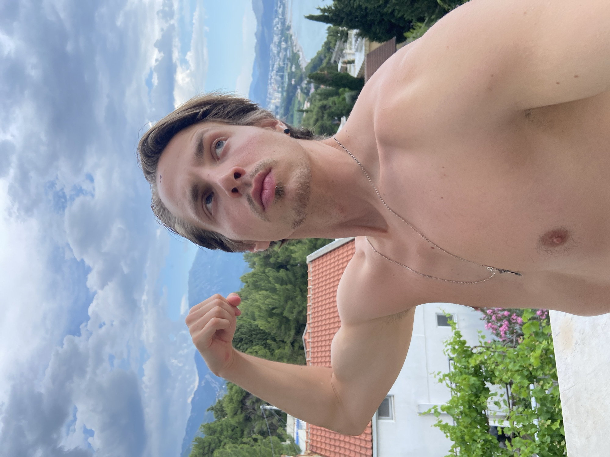 A light-skinned, muscular man stands outdoors in a partially developed location in Šušanj, Montenegro. The man is shirtless, wearing a silver chain necklace. His hair is light brown. He is flexing his left bicep. The background includes an overcast sky, a view of a distant city along the coastline, green trees and shrubs, and a tile-roofed building. The location is elevated.