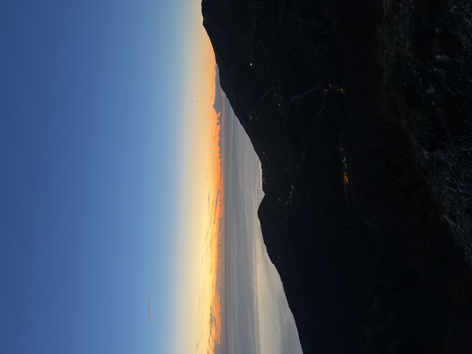 A twilight scene showcases a sharp, dark mountain silhouette against a vibrant, layered sky. The sky transitions from deep blue to orange and yellow, with hints of clouds at the horizon. Small, scattered lights dot the mountainside, suggesting a populated area or path. The mountain's textured surface blends into darkness, contrasting the bright sky.