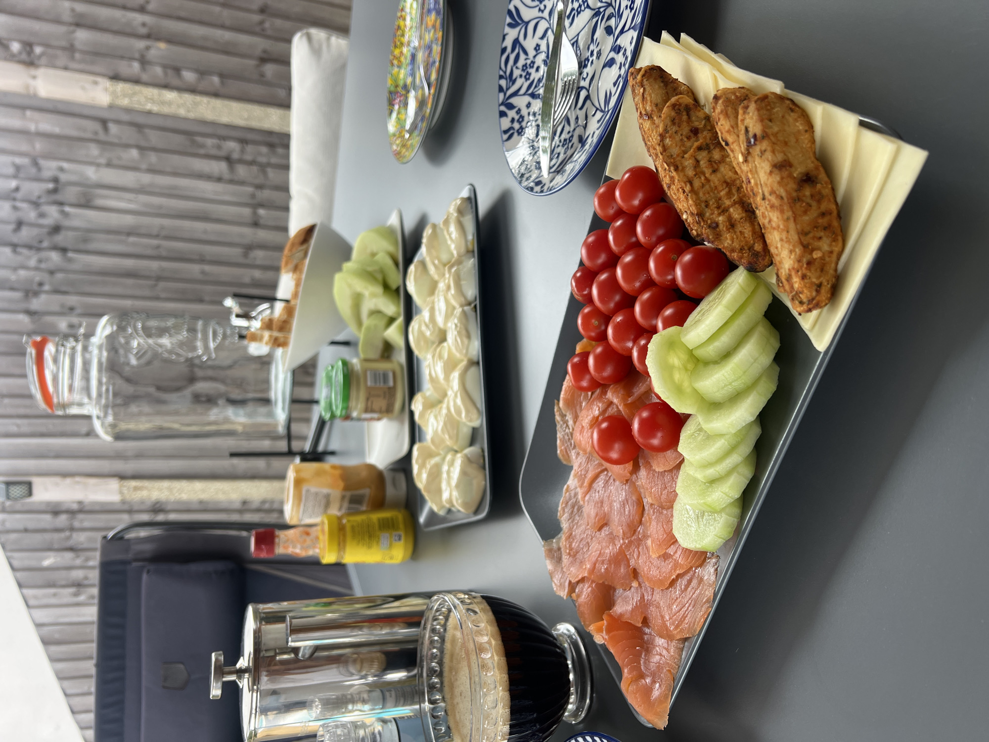 A breakfast spread is laid out on a dark grey table, outdoors, with a blurred wooden fence in the background. The table is laden with a variety of food items on grey platters and a glass bowl. In the foreground, a rectangular grey platter holds thinly sliced smoked salmon arranged in a fanned pattern. Next to the salmon are several halved cherry tomatoes and sliced cucumber rounds. To the right of the platter are two pieces of what appear to be grilled or fried chicken strips, resting on slices of cheese. A glass bowl to the left contains a dark liquid, likely coffee, with foam on top. Behind the main platter, another grey rectangular platter holds what looks like deviled eggs topped with a creamy sauce. To the right of this, a small white bowl contains sliced bread. Several jars, including one yellow and one orange, are visible in the middle ground. A large glass dispenser with a red lid stands prominently in the background. Two decorative plates, one with a blue and white pattern and another with a colorful floral design, are placed on the table further back. A metal French press coffee maker is also visible to the left. The setting suggests a casual outdoor meal, possibly a breakfast or brunch. The specific location in Guildford, United Kingdom cannot be determined from the provided image.