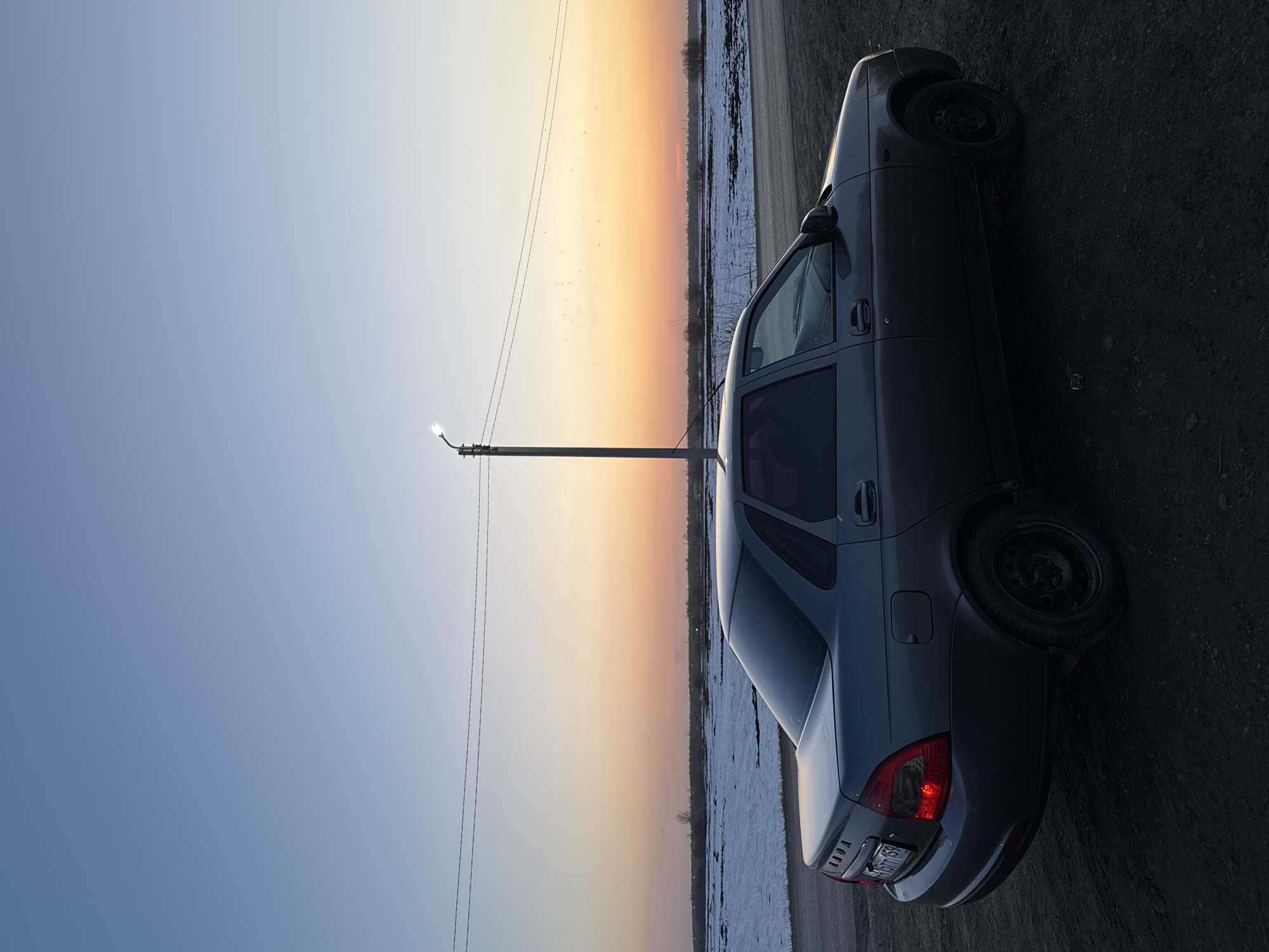A grey Lada sedan is parked on a roadside near Кондратово, Russia. A utility pole stands adjacent to the vehicle, against a backdrop of a sky gradient transitioning from blue to orange, suggesting a sunset or sunrise. A field is visible in the background, partially covered in snow. The road surface appears to be asphalt or gravel. The car is facing away from the camera, revealing its rear lights and license plate.