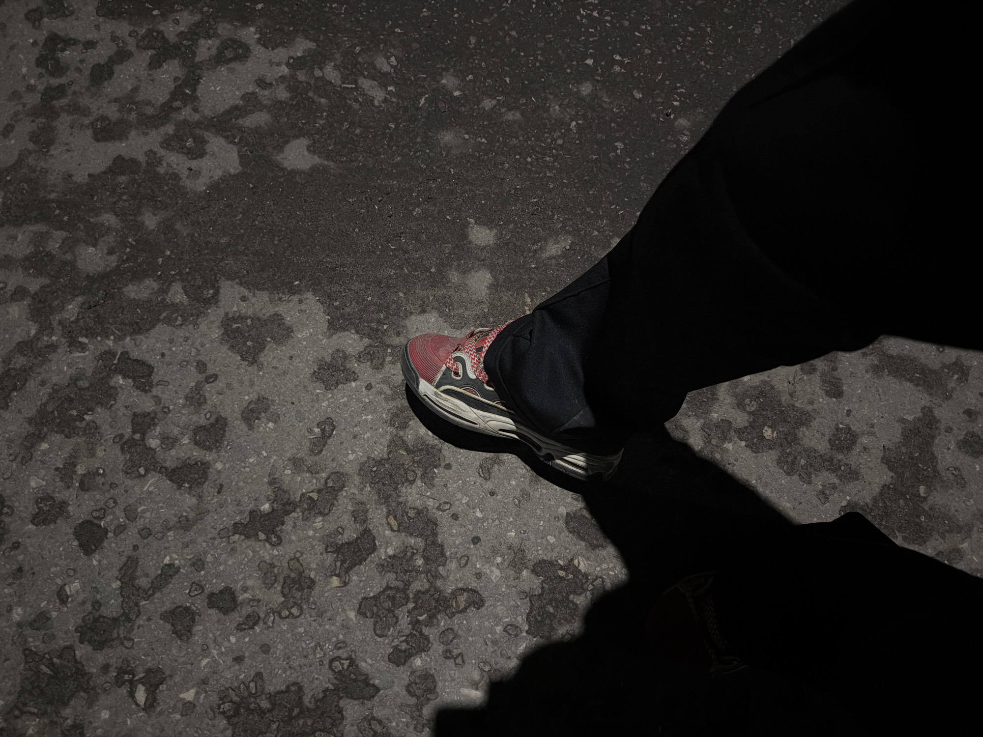 The image shows a person's legs and feet wearing black pants and athletic shoes with red and white accents, standing on a textured asphalt surface. The asphalt has a mottled appearance with areas of darker discoloration and lighter aggregate. A strong shadow is cast to the left of the frame, obscuring much of the ground and partially covering the person's legs. The lighting suggests it is either early morning or late evening. There is no information in the image to suggest the specific location within Nizhny Novgorod, Russia.