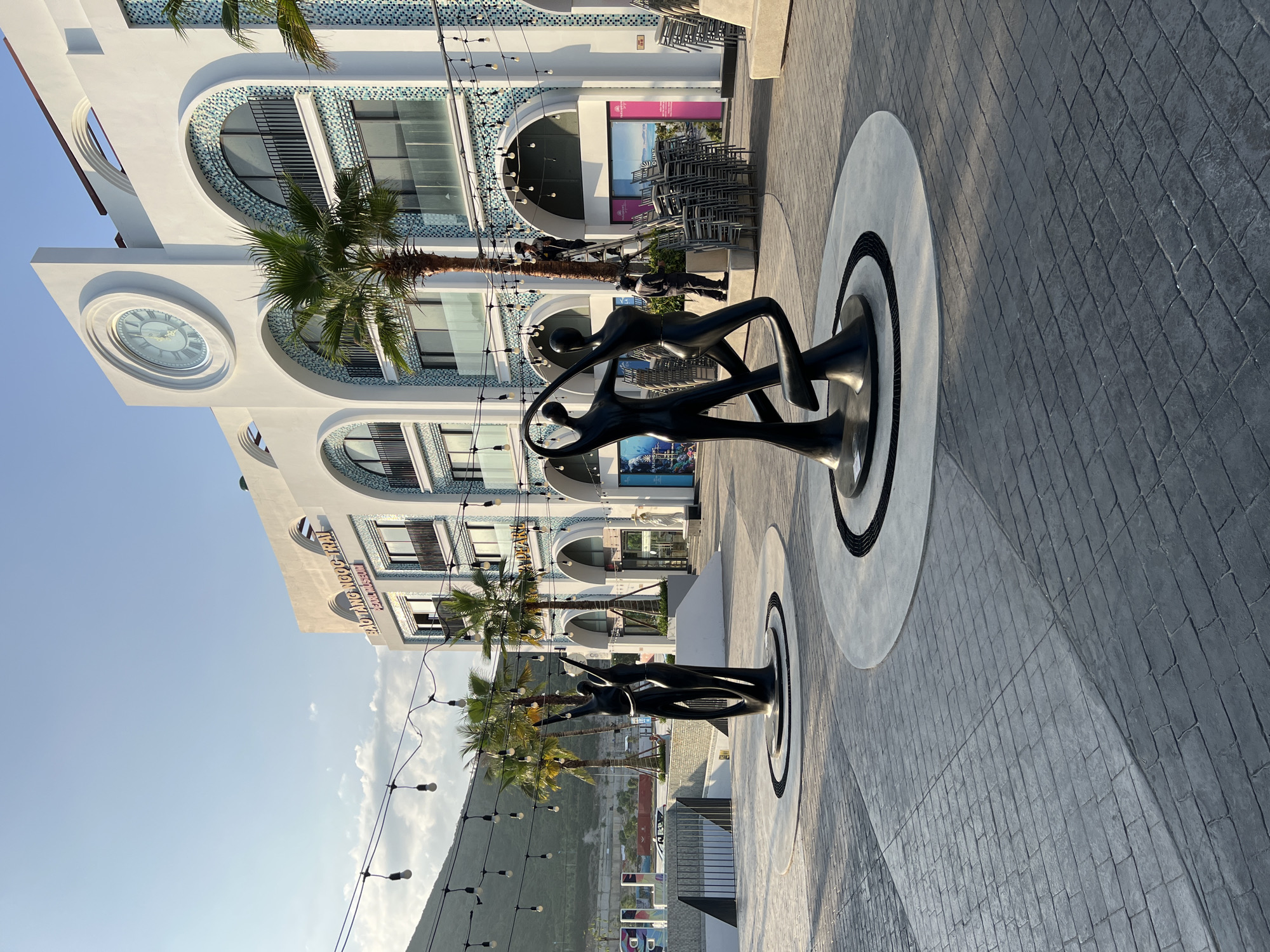 In Nha Trang, Vietnam, an outdoor plaza features two abstract black sculptures of human figures in a dynamic, dancing pose. The sculptures stand on circular bases within a paved area. Behind them, a modern white building with arched windows and decorative blue and white tile accents rises, topped with a large circular clock. Palm trees are scattered throughout the scene. Strings of light bulbs are strung across the plaza, illuminating the area. In the background, a hilly landscape is visible under a clear sky with scattered clouds. The ground is paved with a textured pattern resembling bricks. A few people are visible, including one on a ladder near the building and another standing on the ground, possibly working on the lights or preparing for an event. Stacked chairs are visible near the building, suggesting a temporary setup or storage. The overall scene depicts a public space in Nha Trang, Vietnam, likely designed for leisure or events.