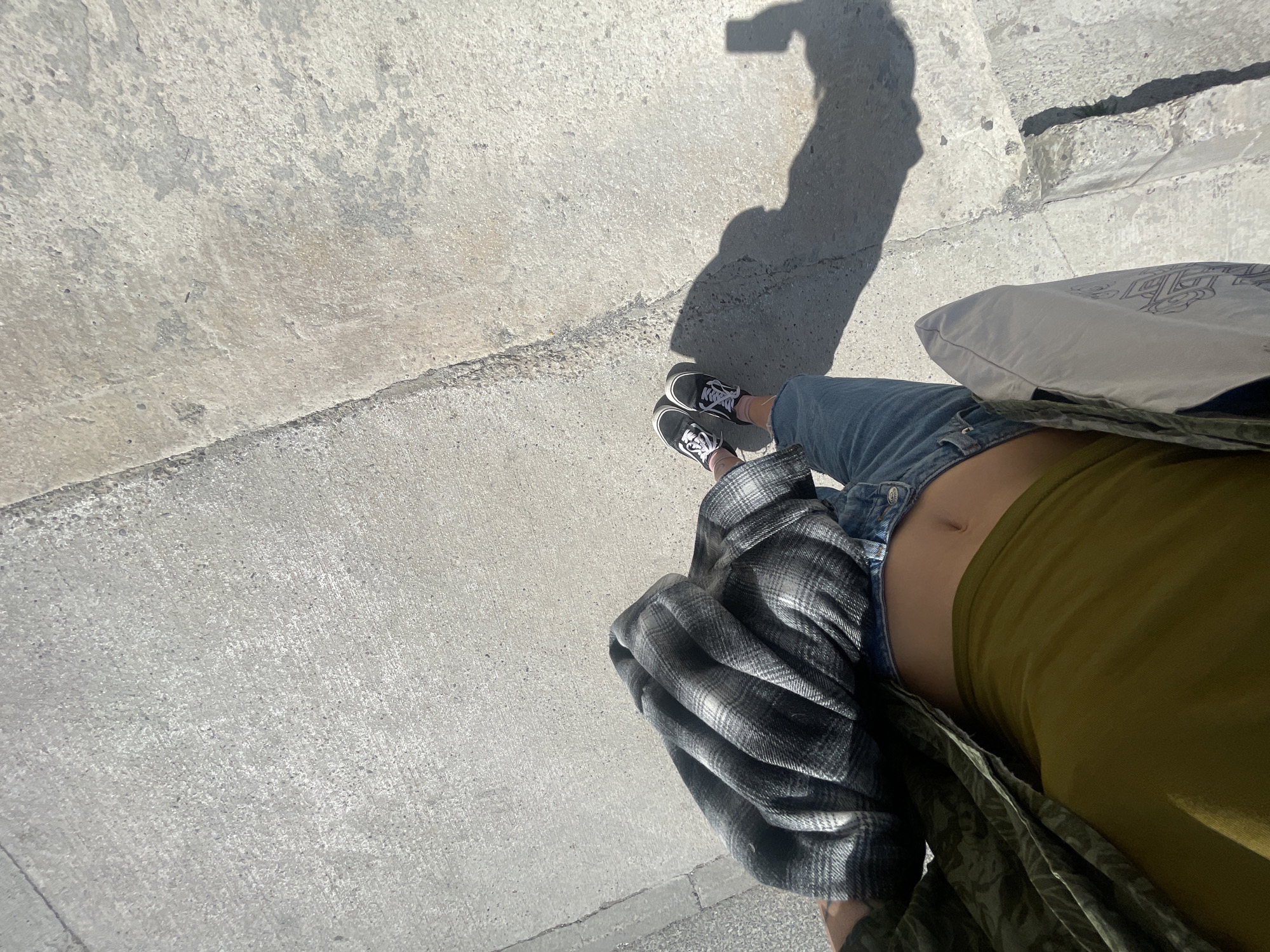 The image shows a person's legs and torso from an overhead perspective. The person is standing on a concrete surface, likely a sidewalk, as indicated by the concrete slabs. They are wearing black and white sneakers, blue jeans, and a green tank top. A gray plaid shirt is draped around their waist. A white tote bag is partially visible on the right side of the frame. The shadow of the person holding the camera is cast on the concrete surface. The location is Boreti, Montenegro, based on external information.
