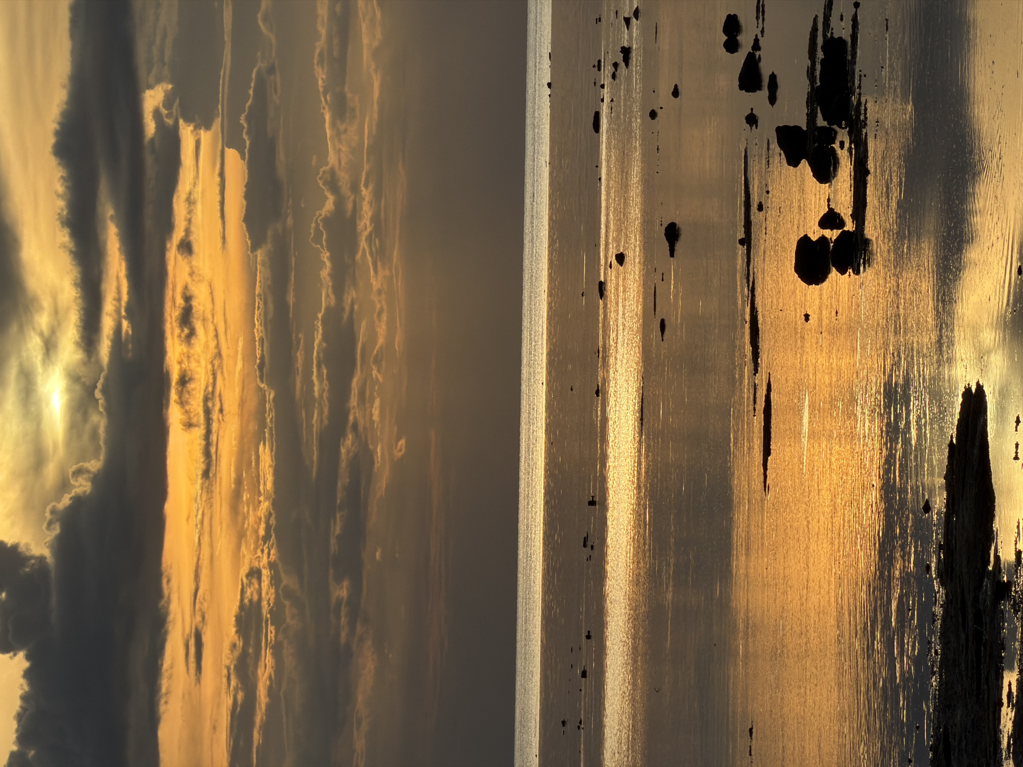 This is a diptych photograph taken at Baan Plai Laem, Thailand. The left panel depicts a dramatic sky filled with elongated clouds, tinged with the warm, golden hues of sunset or sunrise. The sun's light breaks through some of the clouds, creating bright streaks against darker formations. The right panel shows a body of water, likely the sea, reflecting the sky's golden light. Jagged, dark shapes, possibly rocks or debris, are silhouetted against the reflective water. The overall mood is serene and contemplative, capturing the natural beauty of a coastal landscape at dusk or dawn. The lighting suggests it is either early morning or late evening.