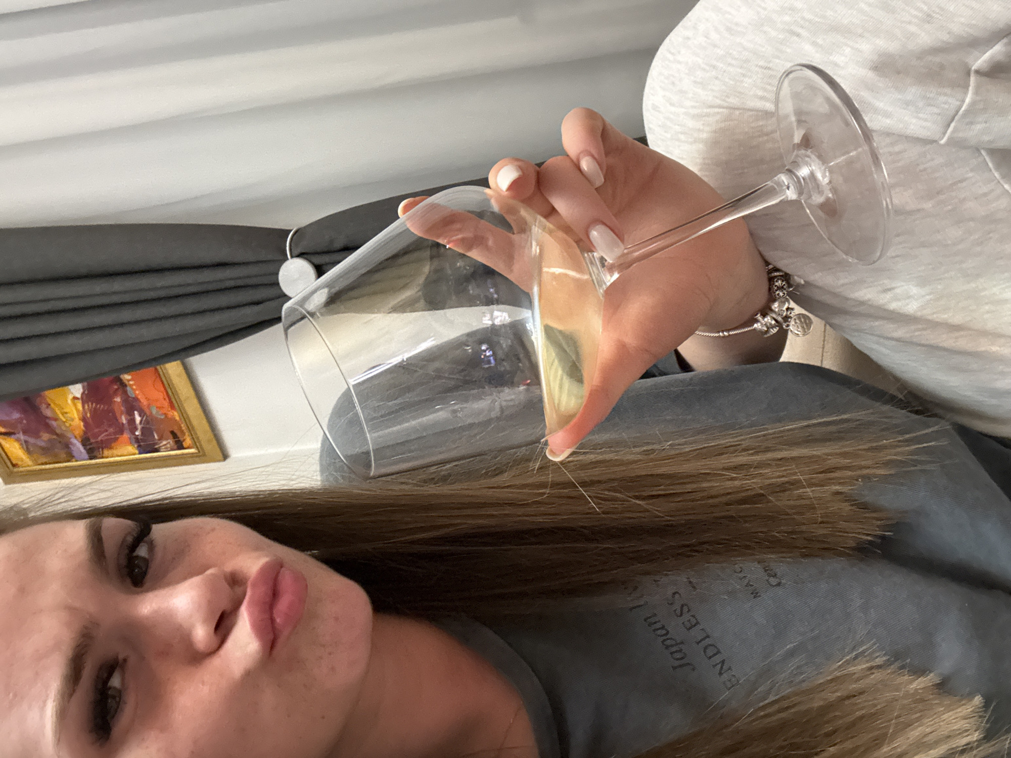 A woman with brown hair and full lips is holding a wine glass filled with liquid. She is wearing a gray t-shirt with the word "Japan" visible. The background includes a painting with a gold frame and a gray curtain with a white rod. The location is likely indoors, but the city of Plovdiv, Bulgaria, cannot be determined from the image.