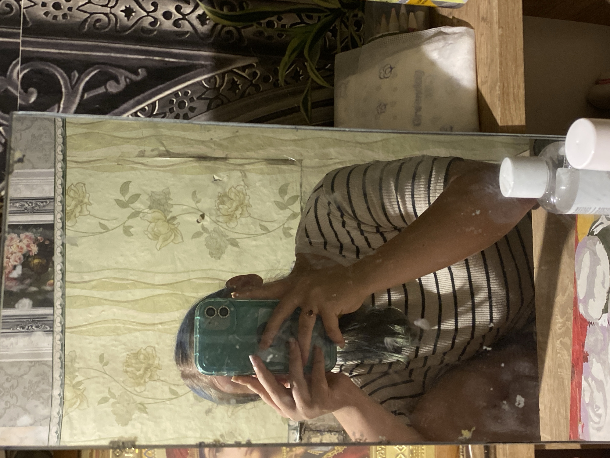 A person is taking a mirror selfie in a room. The person is wearing a striped t-shirt and holding a teal smartphone. The background of the room is decorated with floral wallpaper. On a shelf to the right of the mirror, there are some personal care products and a houseplant. There is no information about the location in the city of Nova Praha, Ukraine.