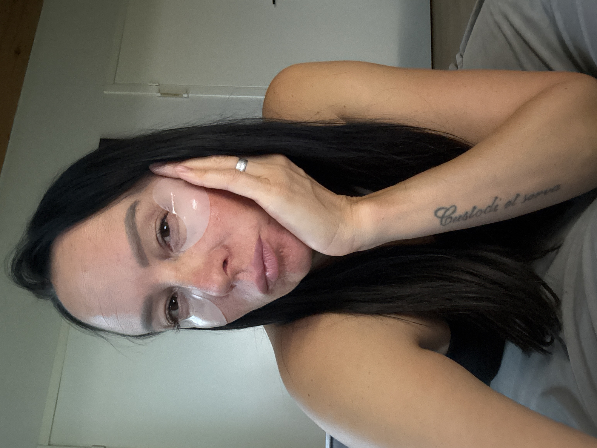The image shows a close-up, high-angle shot of a woman with long, dark hair. She is wearing eye patches under her eyes and has a ring on her right ring finger. Her right arm is resting on her cheek. A tattoo is visible on her left forearm. The background appears to be an indoor setting with a wall and a door. There is no indication of the city of Chaikovsky, Russia, or any specific city in the image.
