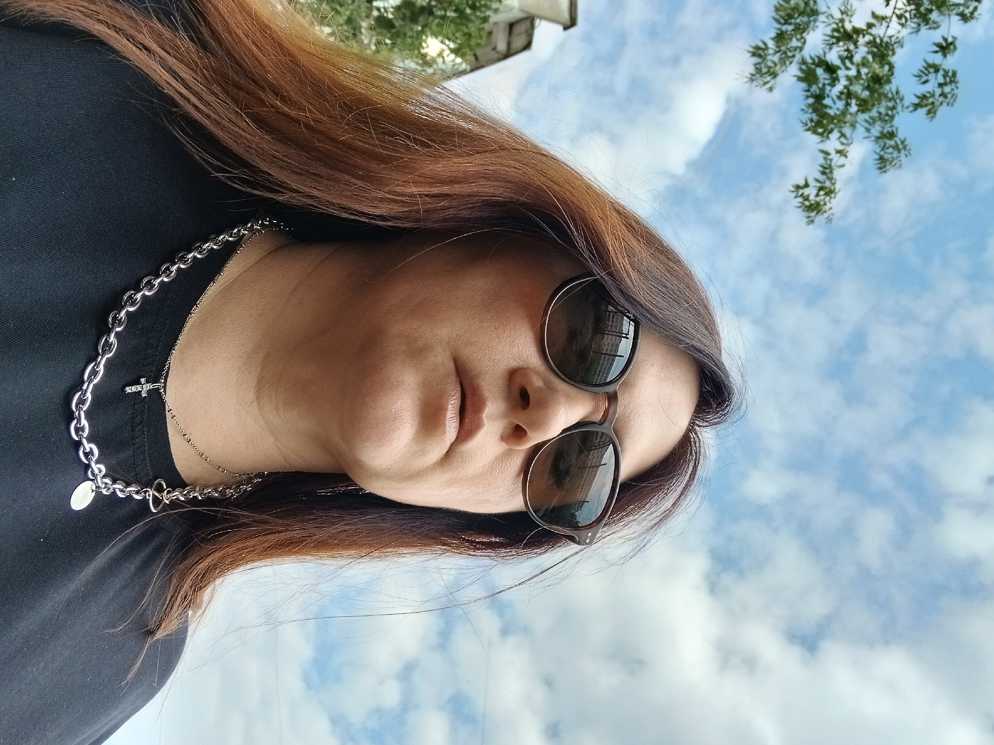 A low-angle, medium shot shows a woman wearing sunglasses and a black t-shirt, with her head tilted slightly upwards. She has long, auburn hair that frames her face and falls over her shoulders. She is wearing a silver chain necklace with a cross pendant. The background consists of a blue sky with white clouds, and a tree with green leaves is visible in the upper left corner. The reflection in her sunglasses shows buildings and the sky. The scene appears to be outdoors, possibly in an urban environment.