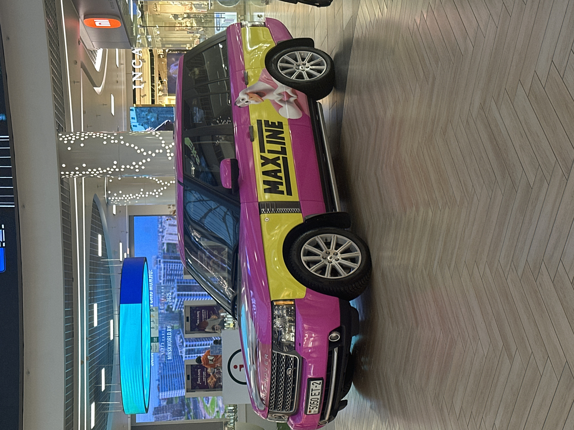 This is a photograph of a brightly colored Range Rover parked inside a modern, well-lit shopping mall in Minsk, Belarus. The vehicle is predominantly pink with yellow accents and features large decals on its side that read "MAXLINE" and depict a cartoonish goose character. The background includes architectural elements of the mall, such as illuminated pillars and signage for shops like "INCA" and "Mi." A large, oval-shaped digital display showing cityscapes and text in Cyrillic characters is visible above the vehicle. The floor of the mall is a polished wooden surface laid in a herringbone pattern. The lighting suggests an indoor commercial environment. The license plate on the vehicle reads "5050 ET-2."