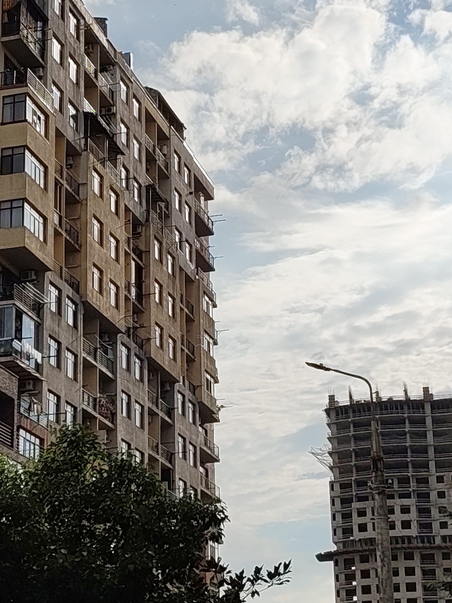 Two buildings dominate the frame against a cloudy sky. The building on the left is a tall, multi-story apartment complex with numerous balconies and windows. Its facade is a mixture of beige and grey concrete, showing signs of age. Some balconies have railings, while others are open. A corner of this building features a glass-enclosed section on a lower floor. Trees with dense green foliage are visible in the foreground on the left, partially obscuring the lower floors of the apartment building.
To the right, a different building is under construction, appearing as a skeletal structure of concrete floors and pillars. A street lamp with a dark fixture is positioned in front of this unfinished building. The sky above is filled with scattered white clouds against a pale blue background. The scene suggests an urban environment, likely Batumi, Georgia, given the architectural style and the presence of both completed and under-construction buildings. There are no visible people in the image. The overall impression is one of urban development and everyday life.