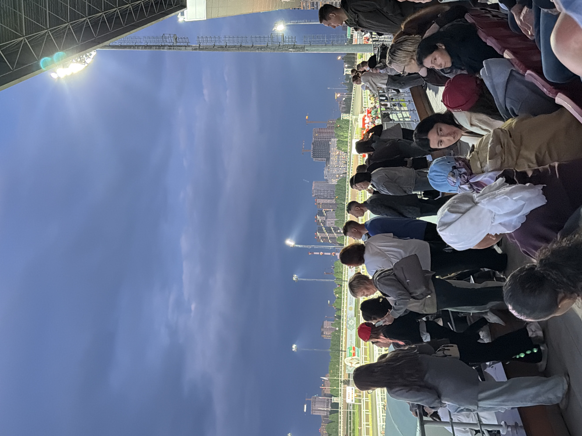 A group of people are standing and sitting in bleachers overlooking a race track. The sky is dark and cloudy, with stadium lights illuminating the track. Several buildings are visible in the background, suggesting an urban setting. The people are dressed in casual clothing, and some are holding phones, possibly taking pictures or videos. The overall atmosphere appears to be one of a sporting event or outdoor gathering.