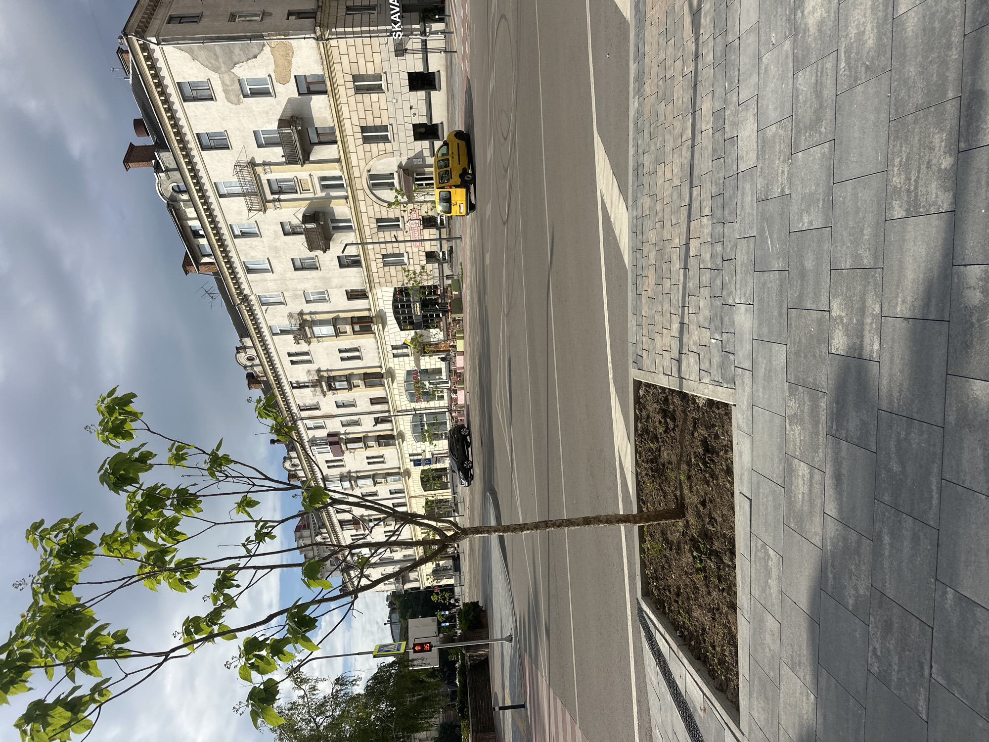 The image captures a street scene in Ternopil, Ukraine, on a cloudy day. In the foreground, a young tree stands in a newly planted square surrounded by modern grey paving stones. The street is paved and marked with white lines. Across the street, a multi-story building with an aged facade dominates the background. The building features several balconies and a row of windows. A yellow van is visible turning in the street, with tire marks suggesting recent maneuvering. A black car is parked further down the street. A traffic light showing "35" is visible on the left side of the image, along with a pedestrian crossing. The overall impression is of an urban environment experiencing a period of modernization, as indicated by the new paving.
