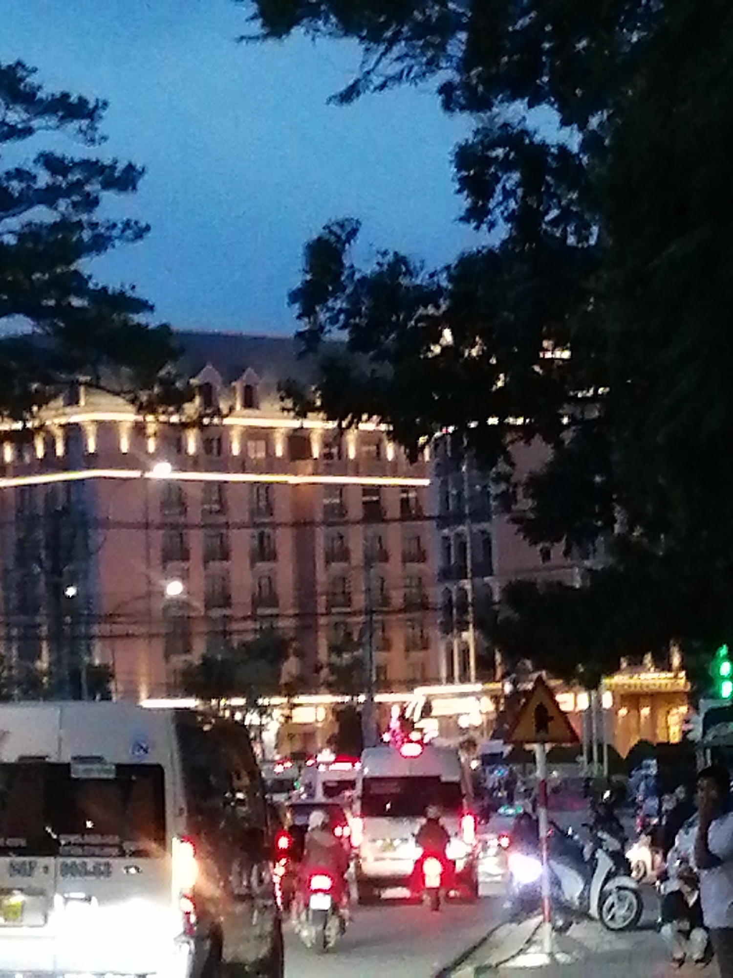 The image depicts a street scene at dusk or night in Lâm Viên - Đà Lạt, Vietnam. A multi-story building with illuminated windows and decorative lighting is visible in the background. Trees with dark foliage frame the upper portion of the image. In the foreground, traffic is present, including a white van with visible license plate markings and other vehicles, including motorcycles, with their lights on. Several individuals are visible on the street, some on motorcycles and others standing. A yellow triangular traffic sign with a black symbol is mounted on a pole. The overall lighting suggests an urban environment during low-light conditions.