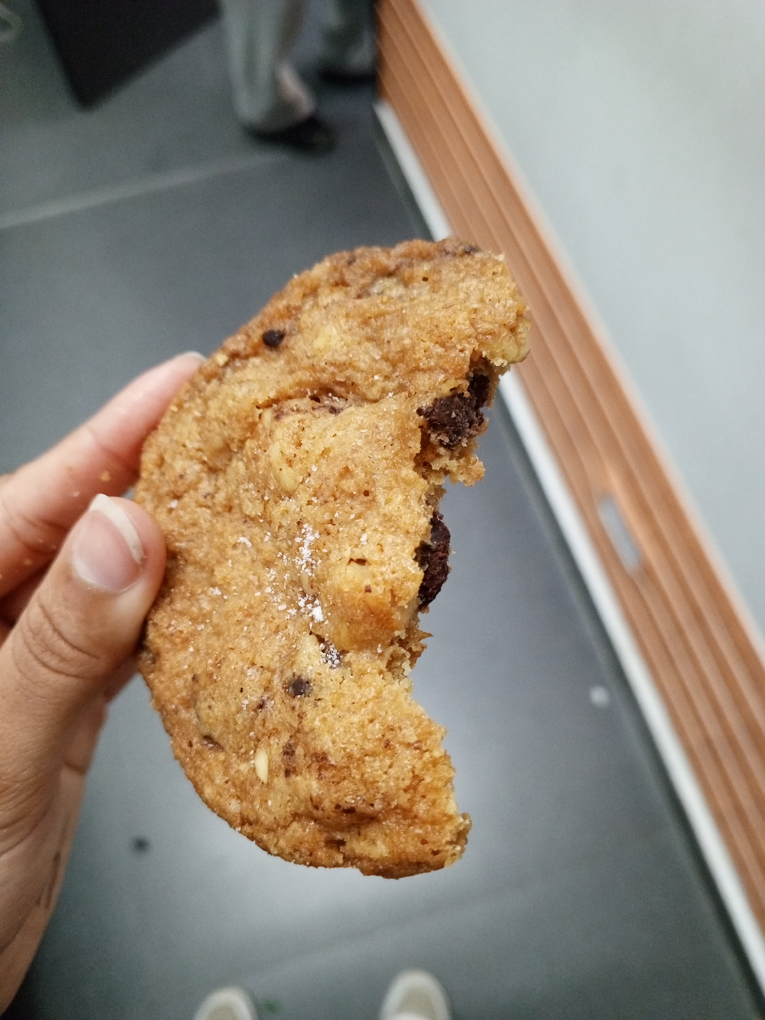 A partially eaten chocolate chip cookie is held in a hand with a gray fingernail. The cookie has a light brown color with dark brown chocolate chips visible. A bite has been taken out of the cookie. In the background, there is a gray floor and a wall with alternating white and brown stripes. There is also a glimpse of two people's feet wearing shoes in the background. The location is unknown and not Pampatar, Venezuela.