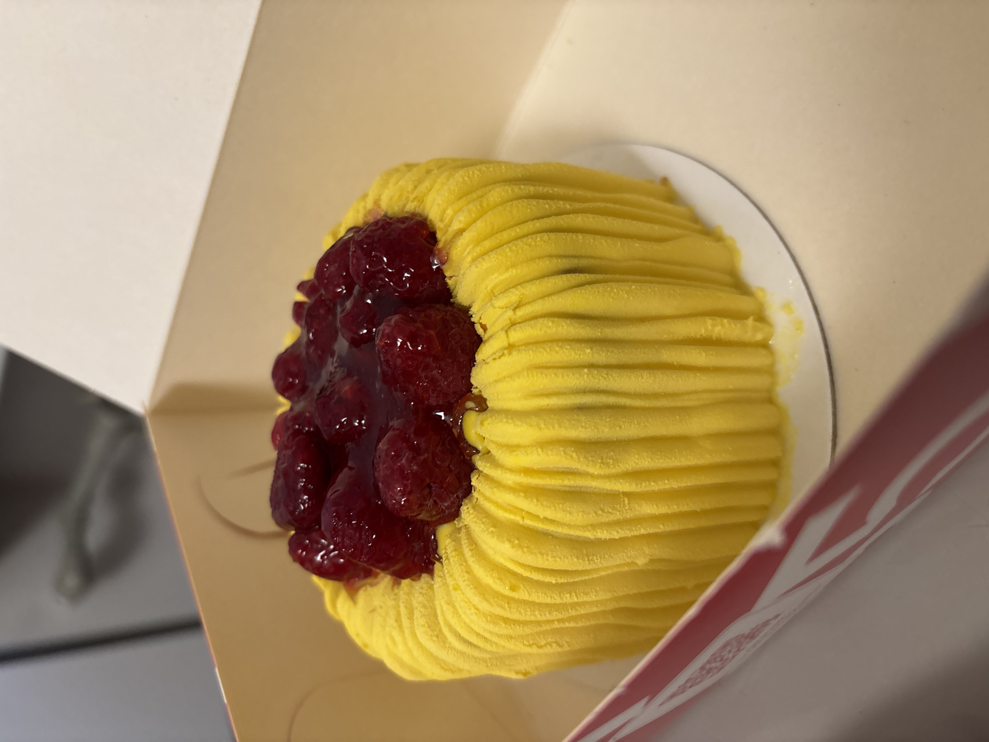 A close-up shot shows a dessert resembling a Mont Blanc pastry inside a box. The pastry is on a white plate. It features a swirl of bright yellow frosting resembling thin strands covering a dome-shaped base. Half of the pastry is topped with a generous heap of raspberries, covered in a transparent, slightly viscous glaze or sauce. The box is a light cream color with a red and white pattern on the inside. The background is out of focus, but parts of what look like a counter can be seen. There is no indication of the location being Saint Petersburg, Russia.