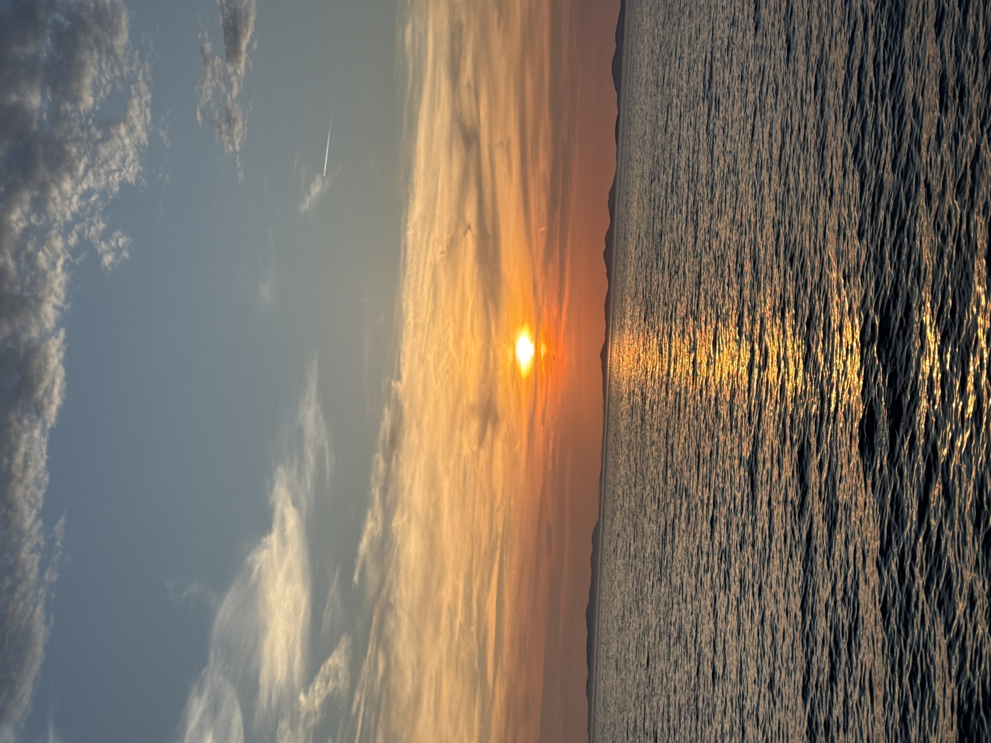 The image depicts a sunset over the Adriatic Sea, viewed from the vicinity of Primošten, Croatia. The sun, a bright orb, is setting on the horizon, casting a warm, orange glow across the sky and reflecting on the water. The sky transitions from a pale blue at the top to vibrant shades of orange and yellow closer to the horizon, with scattered wispy clouds. In the distance, a silhouetted chain of islands forms a low, undulating line against the colorful sky. The sea in the foreground is dark blue, with ripples catching the sunlight to create a shimmering path. The overall scene is serene and captures the natural beauty of a coastal sunset.