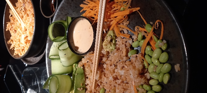 The image captures a close-up, slightly overhead view of two food dishes, likely in an indoor, dimly lit restaurant setting, possibly during the evening.
The primary focus is a dark, round bowl containing an Asian-inspired meal, resembling a poke bowl or rice bowl. It features a bed of light brown rice, topped with a vibrant array of ingredients. On the left, there are several slices of green avocado and thin, curled ribbons of cucumber. In the center, a small black cup holds a creamy, light orange-pink sauce. To the right, bright green edamame beans are scattered among shredded orange carrots. A small dollop of green paste, possibly wasabi or mashed avocado, is visible near the center. A pair of light-colored chopsticks is resting over the main dish, poised for eating.
To the upper left of the main dish, partially visible, is another dark bowl filled with light orange-brown noodles, also with a pair of chopsticks inserted into them.
The background is predominantly dark, suggesting low ambient lighting, possibly from a lamp or overhead fixture, which illuminates the food. A clear glass is partially visible in the bottom left corner, reflecting some light. No discernible text is visible in the image.