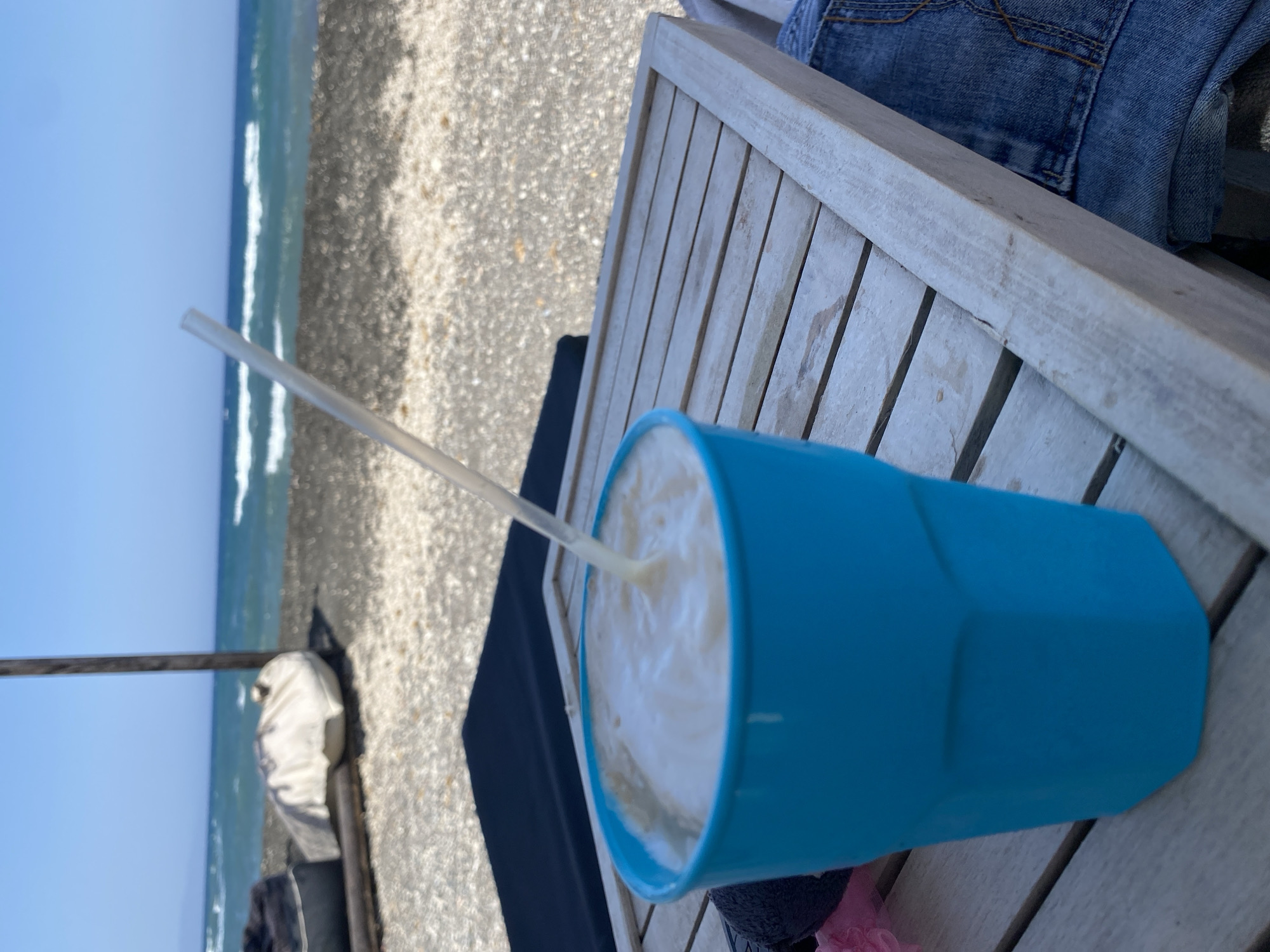 A close-up shot of a bright blue plastic cup filled with a frothy, light-colored beverage sits on a weathered wooden slatted table on a beach. A clear plastic straw extends from the drink and angles towards the top right of the frame. In the background, the sea is visible with gentle waves under a clear blue sky. A dark, lumpy object, possibly a cushion, and a lighter, rounded object covered in white fabric are situated on the pebbly shore behind the table. A wooden pole stands vertically near the lumpy object. Part of a person's denim-clad leg is visible in the lower right corner, suggesting they are seated at the table. The scene is likely at a beachside establishment in Thira Municipal Unit, Greece, given the characteristic Aegean Sea backdrop and pebbly shoreline.
