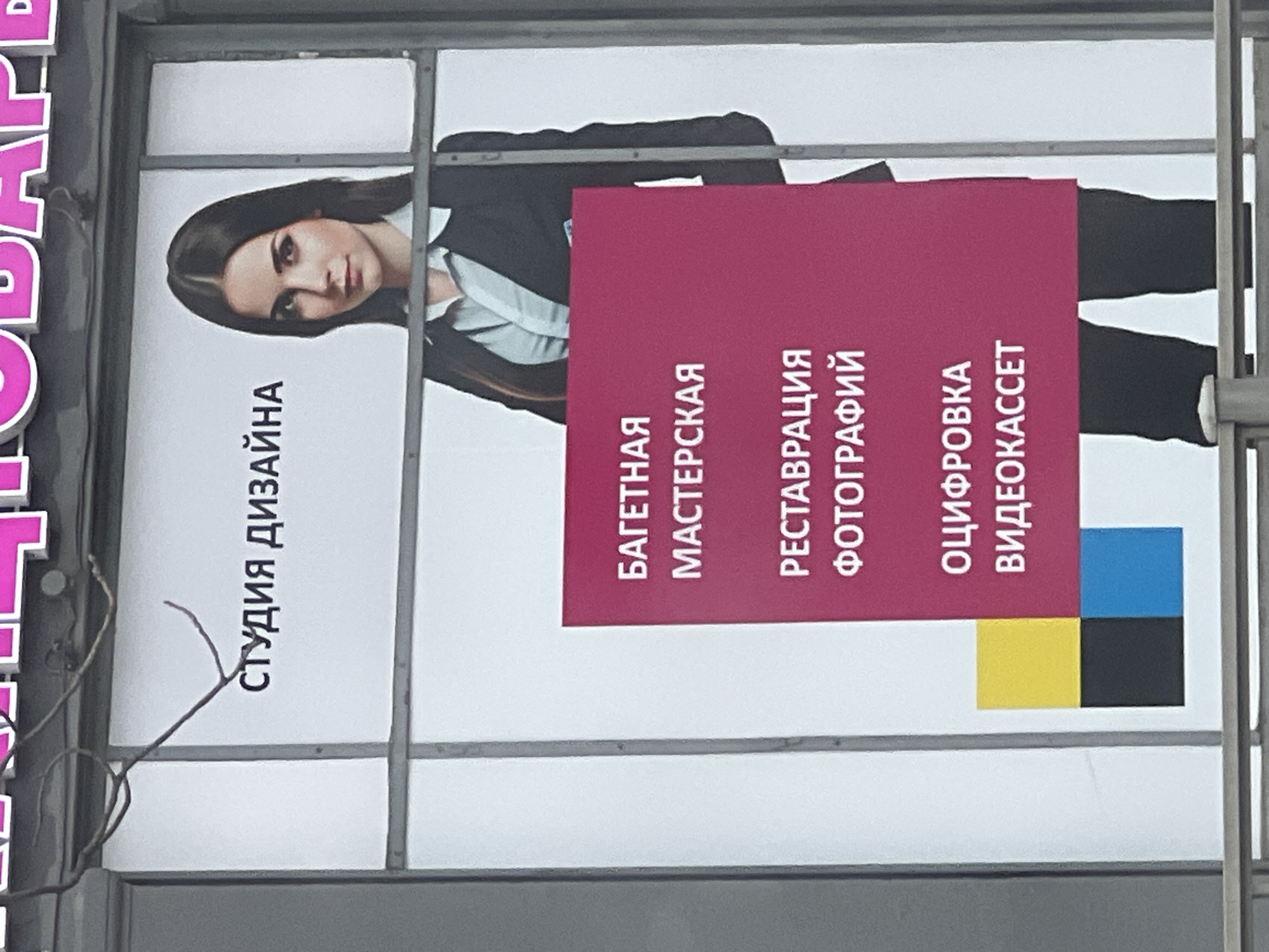 The image displays a large outdoor advertisement or billboard, captured from a slightly upward angle and rotated 90 degrees clockwise. The advertisement features a young woman and text in Russian, set against a plain light background, likely white.
On the left side of the sign (when viewed with the text oriented correctly), a woman with long, dark, straight hair and a neutral expression is depicted. She is wearing a dark suit jacket over a light-colored, possibly white or light blue, collared shirt. Her head, shoulders, and upper torso are visible, looking directly at the viewer.
Overlaying part of the woman's body and extending to the right is a prominent, dark red (maroon or burgundy) rectangular box. This box contains three lines of vertically oriented Russian text (when the image is rotated correctly).
The text visible on the sign includes:
1. To the left of the woman's head, partially obscured by thin branches or wires: "СТУДИЯ ДИЗАЙНА" (STUDIYA DIZAYNA), which translates to "DESIGN STUDIO."
2. Within the dark red rectangular box, listed vertically from top to bottom:
* "БАГЕТНАЯ МАСТЕРСКАЯ" (BAGETNAYA MASTERSKAYA), translating to "FRAMING WORKSHOP."
* "РЕСТАВРАЦИЯ ФОТОГРАФИЙ" (RESTAVRATSIYA FOTOGRAFIY), translating to "PHOTO RESTORATION."
* "ОЦИФРОВКА ВИДЕОКАССЕТ" (OTSIFROVKA VIDEOKASSET), translating to "VIDEOCASSETTE DIGITIZATION" or "DIGITIZING VIDEO TAPES."
In the bottom right corner of the dark red box, there are three colored squares arranged in an L-shape: a bright blue (cyan) square at the top, a yellow square below it, and a black square to the right of the yellow one. These colors suggest a CMYK print color model.
The entire advertisement is enclosed within a sturdy grey metallic frame, with visible horizontal and vertical support bars segmenting the sign into panels. On the far left edge of the image, a small portion of another sign with large pink letters (partially visible "P" and "Б") can be seen. Thin, dark, gnarled branches or wires obscure a small part of the "СТУДИЯ ДИЗАЙНА" text on the left side of the main sign. The background behind the sign appears to be a uniform light color, possibly an overcast sky or a distant building.