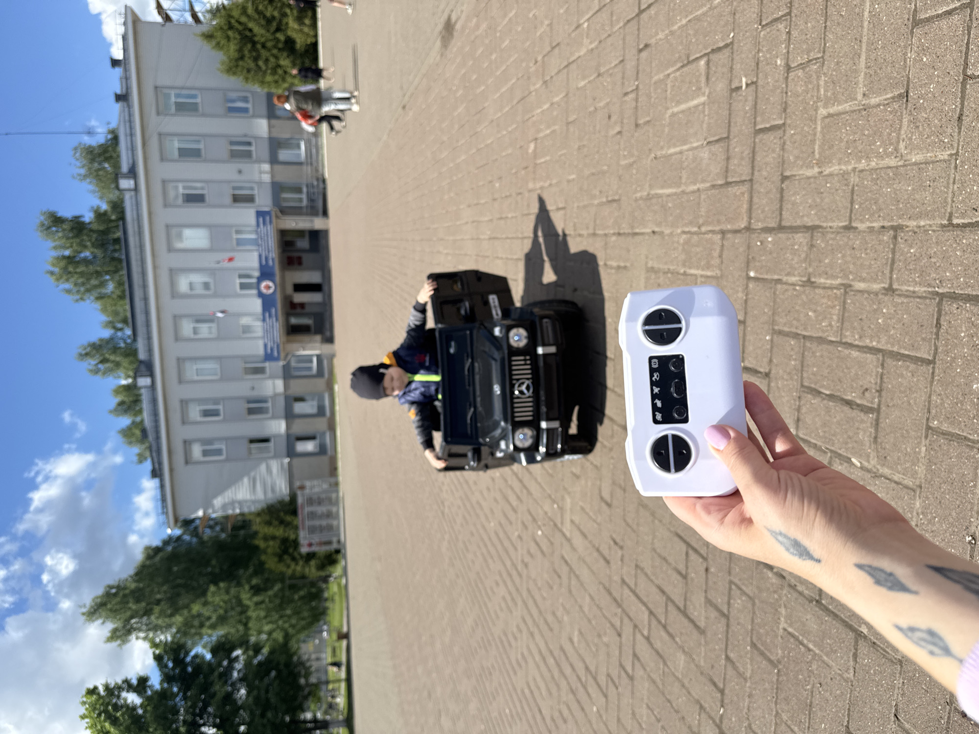 A slightly skewed outdoor shot reveals a brick-paved square in Zav'yalovo, Russia, on a sunny day. A child sits in a miniature black Mercedes G-Wagon, positioned toward the viewer. A white remote control is held in the foreground by a female hand featuring feather tattoos on the arm and manicured fingernails. The remote is presumably used to operate the toy car. The building in the background is a light grey, multi-story structure with visible windows and architectural details. Other people are visible walking in the background near the building. Trees flank the building on either side. The sky is blue with sparse clouds. The shadow of the car and the person standing taking the picture is cast onto the bricks.