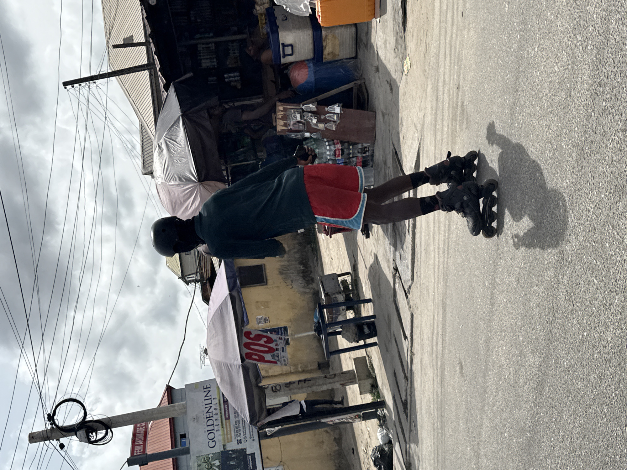 The image captures an outdoor street scene in Ado, Nigeria, under bright, overcast daylight, evidenced by distinct shadows. The scene is viewed from slightly behind a person on rollerblades, looking towards a row of street stalls.
The central subject is a person wearing a dark long-sleeved top, red shorts with light blue trim, dark socks, and a black helmet, standing on black inline rollerblades. Their shadow is prominently cast on the textured, paved ground, which appears to be asphalt or concrete.
To the right side of the frame (behind the rollerblader from the perspective of the original photo orientation), several makeshift stalls or small shops line the street. One stall is shaded by a light-colored umbrella. Various goods are displayed for sale, including bottled drinks like Coca-Cola and numerous small plastic bottles, possibly containing medicines or toiletries. Large coolers are also visible.
Overhead, a dense network of electrical or utility wires stretches across the cloudy sky, supported by utility poles. Buildings with corrugated roofs can be seen behind the stalls.
Several signs are visible: a prominent red "POS" (Point of Sale) sign hangs under one of the umbrellas. A large white signboard for "GOLDENLINE SCHOOLS" is also present, with smaller text indicating "PRIMARY," "FEMI AKINBADE CO," and contact numbers. A partial sign bearing "FEMI AKINBADE" is also discernible.
The overall scene portrays daily street commerce and activity, with the rollerblader providing a focal point.