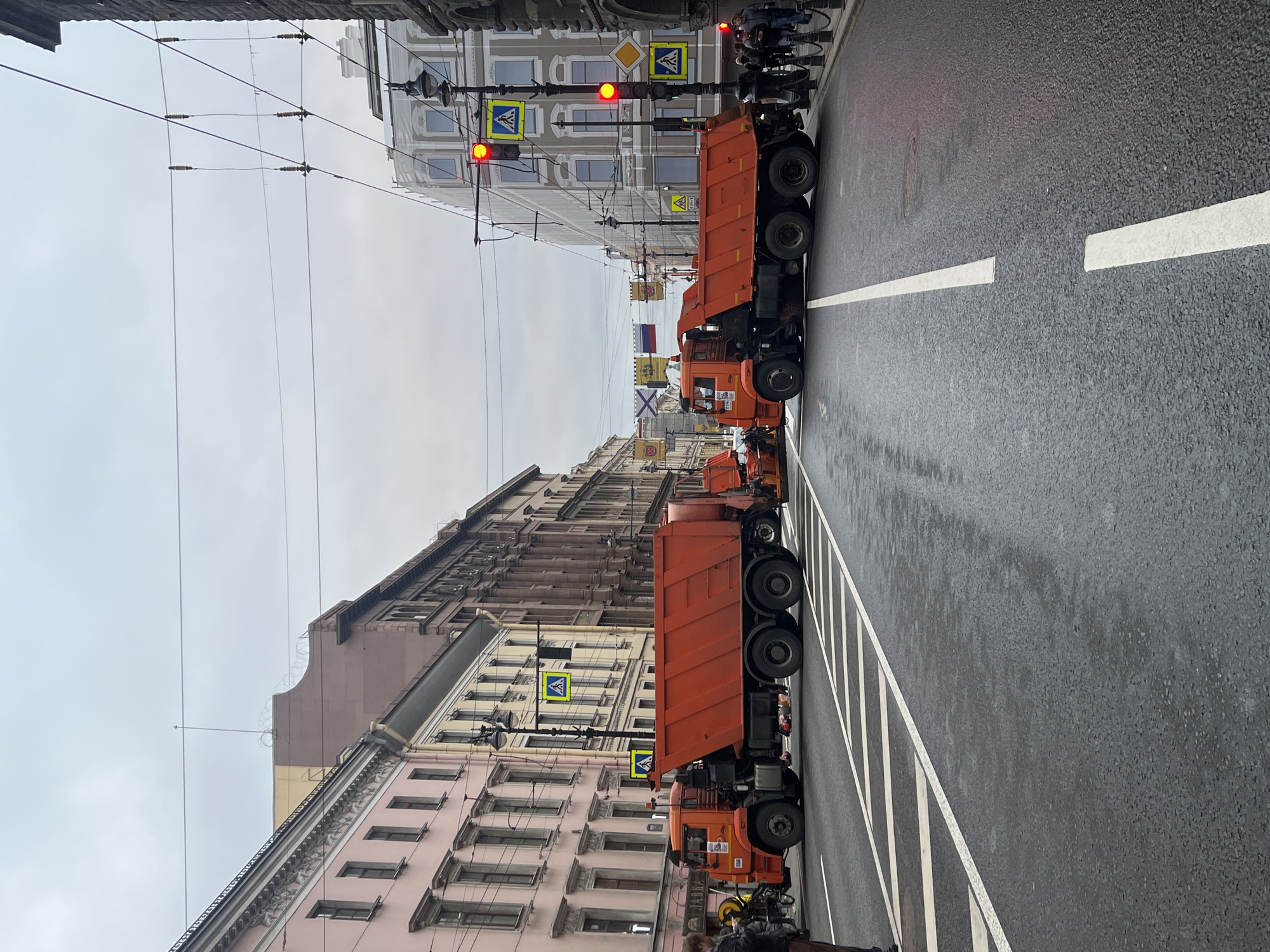 The scene is a street in Saint Petersburg, Russia, likely a central area due to the presence of older, multi-story buildings with detailed architectural facades. A row of orange dump trucks is parked down the center of the street, straddling a solid white center line. The street surface appears wet. Pedestrian crossings are marked with zebra stripes and warning signs. Traffic lights are visible and show red. Overhead tram or trolleybus wires are present. People are visible on the sidewalks near the trucks. Russian flags are visible. The sky is overcast.