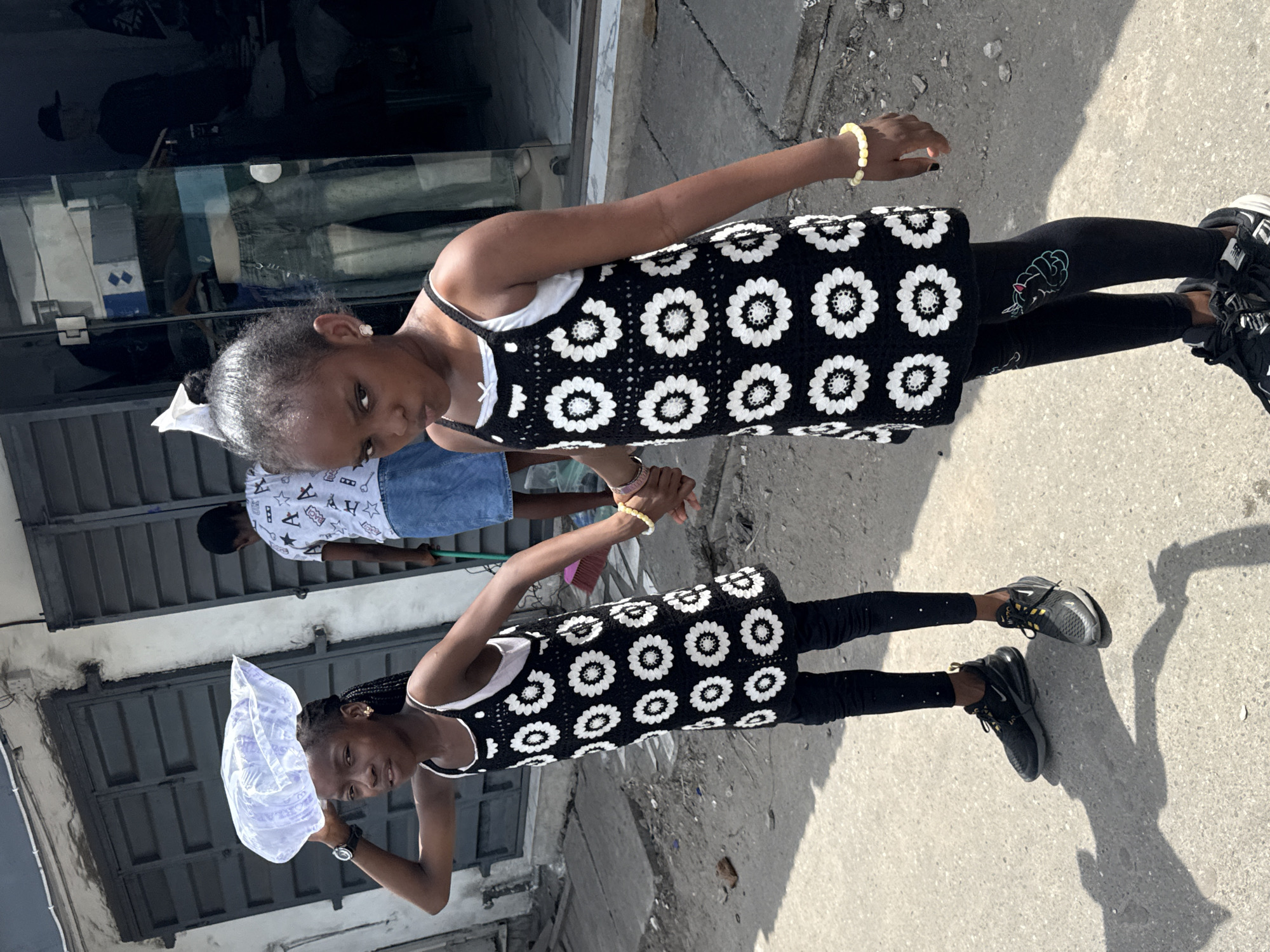Two young girls, likely sisters, stand side-by-side on a concrete sidewalk in an urban environment, possibly Lagos, Nigeria. The girl on the left holds a translucent plastic bag above her head, while the girl on the right holds the other's hand. Both wear black and white floral dresses, leggings, and sneakers. A person wearing a patterned shirt and jeans is partly visible in the background. The scene is outdoors, with buildings and a glass-fronted storefront partially visible in the background. The light suggests it is daytime.