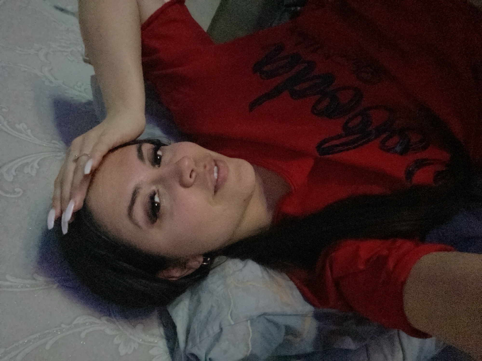 A woman is lying on a bed with a patterned blue and white comforter. She is wearing a red t-shirt with black lettering. Her right hand is resting on her forehead. She has dark hair and fair skin. She is wearing a ring and has long white nails. The location is not specified.