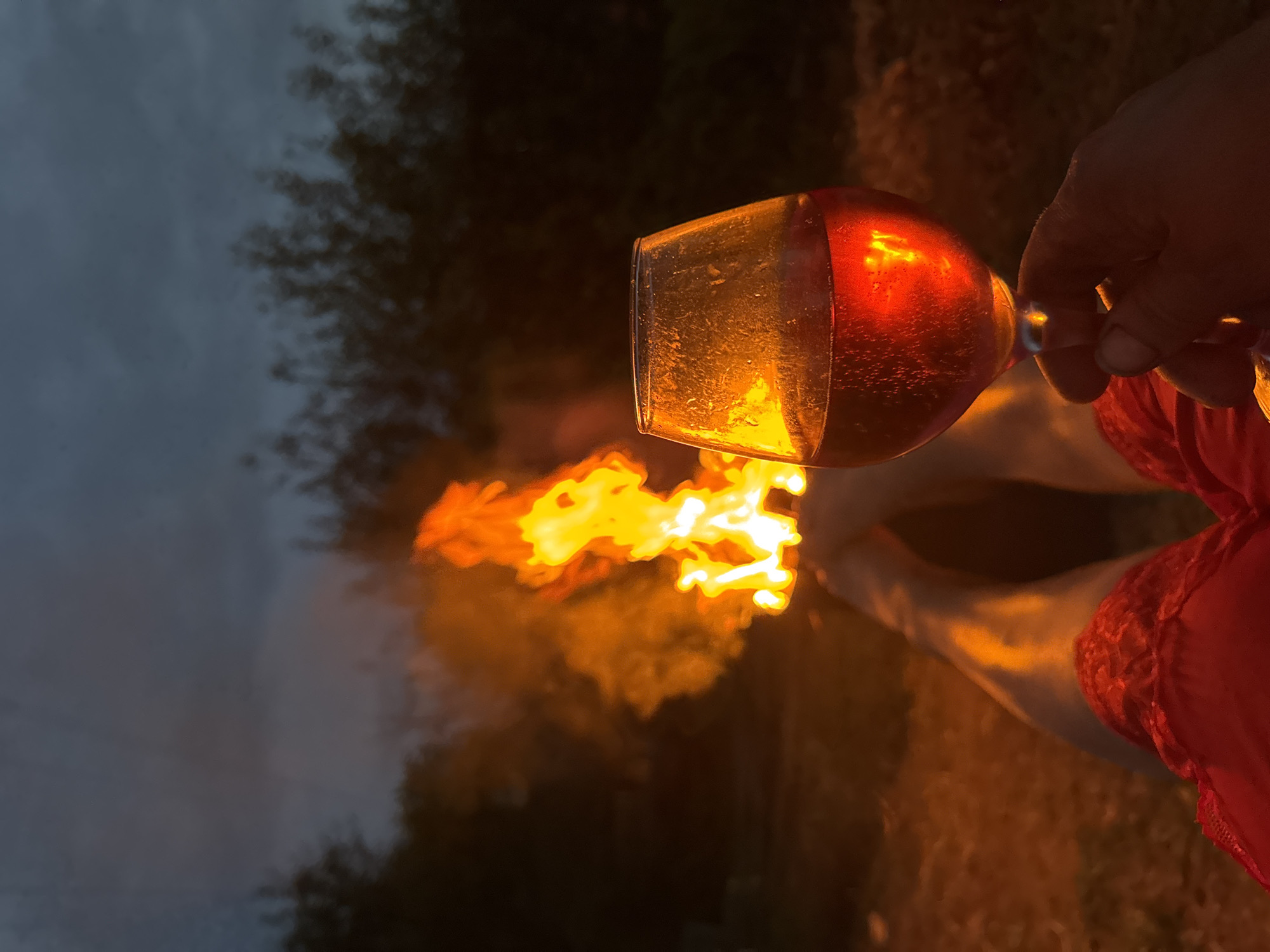 A person's legs, clad in red shorts, are extended towards a crackling bonfire. The flames cast a warm glow on the skin and the surrounding ground, which appears to be covered in dirt and possibly grass. In the foreground, a hand holds a wine glass filled with a reddish liquid, likely wine or a similar beverage. The glass is partially illuminated by the fire, creating an orange hue on its surface. The background is dark, with silhouetted trees against a twilight sky. The overall atmosphere is one of relaxed outdoor leisure, possibly in a rural or semi-rural setting on the outskirts of Chaikovsky, Russia.