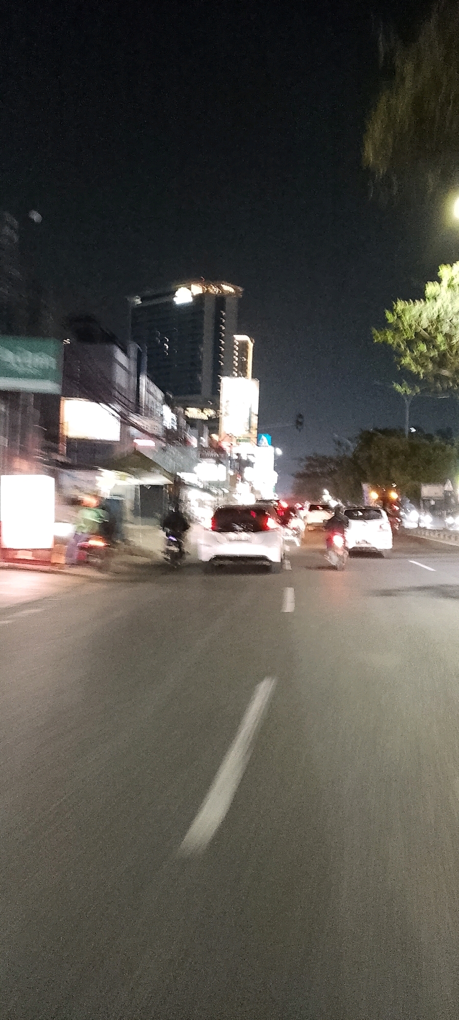 The image depicts a nighttime street scene in Depok, Indonesia, likely on a busy road. A blurred white car is the most prominent subject in the foreground, seen from the rear. To its left, a motorcycle with a rider is also blurred, suggesting movement. Further down the road, other vehicles and streetlights are visible, contributing to the overall impression of a bustling urban environment at night. Tall buildings with illuminated windows can be seen in the background, indicating the presence of commercial or residential structures. The dark sky, punctuated by artificial lights, suggests the photo was taken after sunset. The image's blurriness indicates motion blur, possibly due to the camera or vehicles moving during the exposure.