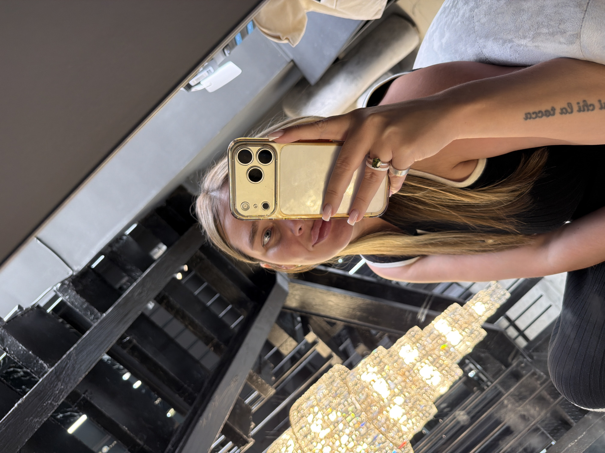 A selfie taken in a location in Moscow, Russia, possibly a commercial building. The subject is a light-skinned woman with blonde hair, holding a gold iPhone in front of her face. She wears several rings on her fingers and has a tattoo on her forearm. Below her is a large, elaborate chandelier and a metal staircase. Other indistinct objects and surfaces are visible in the background.