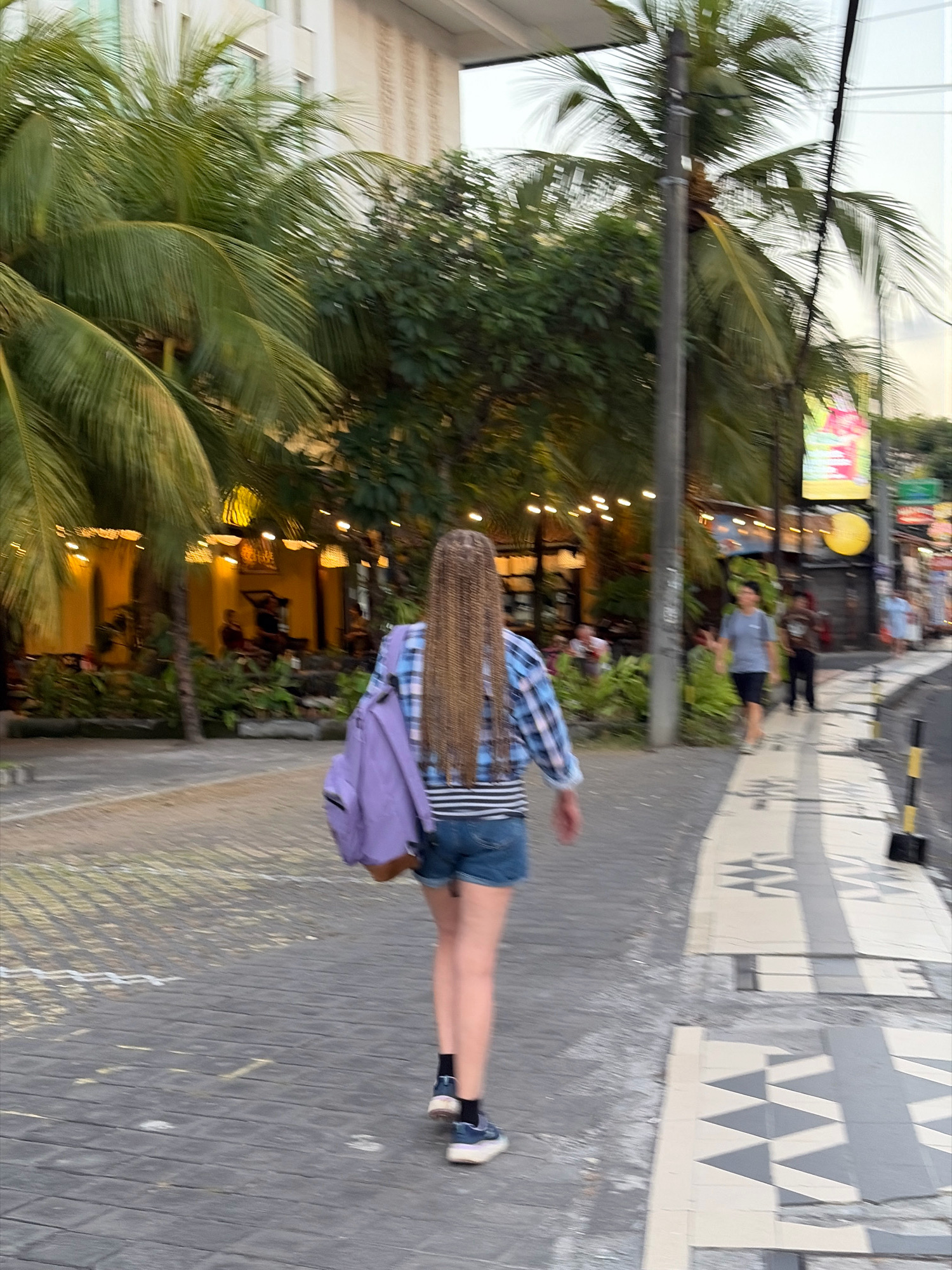 A full-body, rear view of a person with long, braided hair walks away from the camera down a sidewalk in Kuta, Indonesia. The person wears a blue and white plaid shirt, denim shorts, and black socks with blue and white sneakers. A light purple backpack is slung over their left shoulder. The sidewalk is paved with gray and white tiles featuring a geometric pattern. To the left of the sidewalk are palm trees, lush green foliage, and buildings with yellow lighting. To the right of the sidewalk, a street is visible with other pedestrians and a colorful advertisement sign. The lighting suggests it is either early morning or late afternoon.