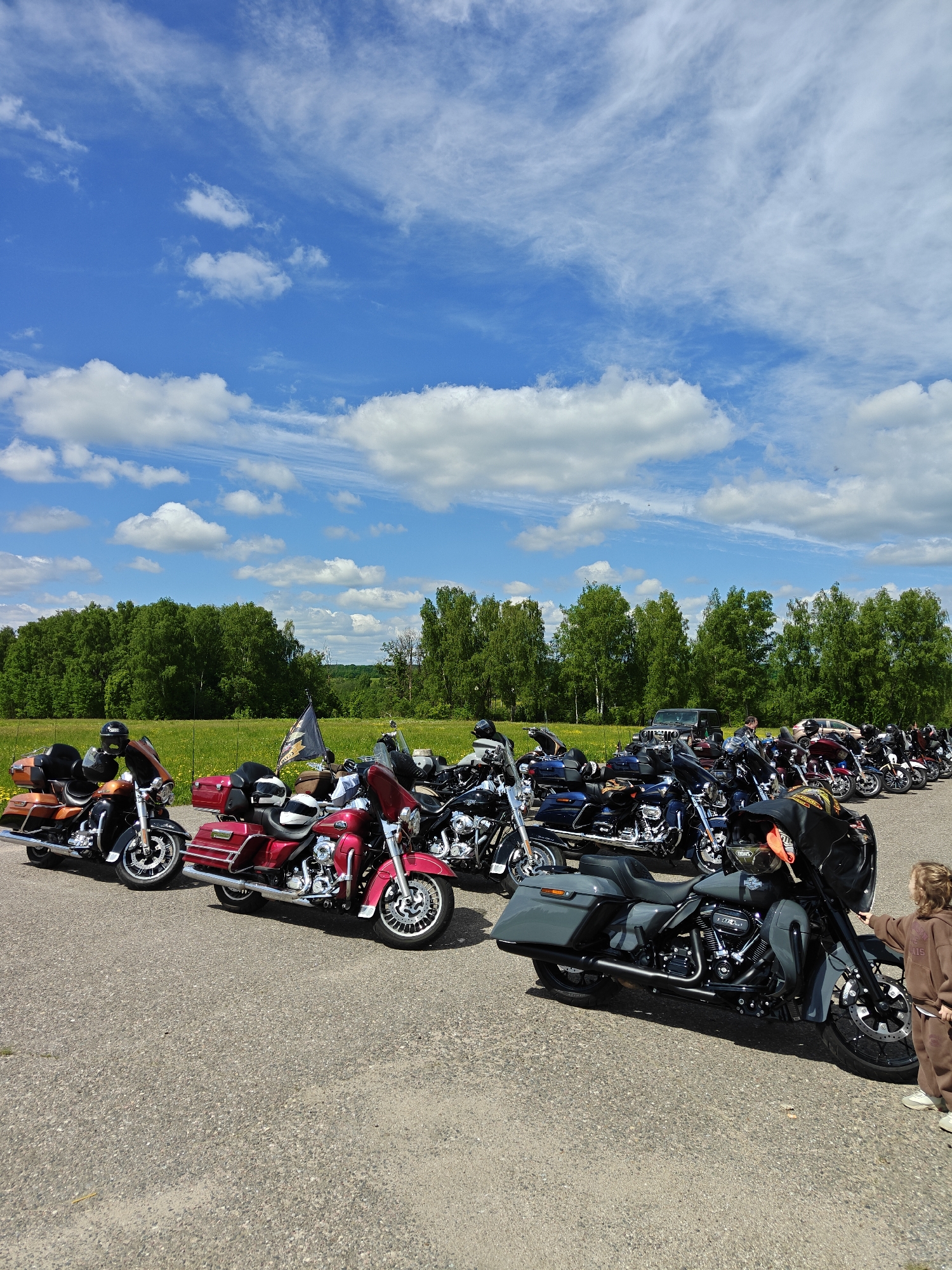 A wide outdoor shot shows a parking lot filled with motorcycles under a blue sky with scattered white clouds. The motorcycles, predominantly Harley-Davidson models, are arranged in rows. To the front-right of the scene stands a child in a brown jumpsuit, reaching out towards a grey motorcycle. The background includes a green field bordered by a line of trees. A dark-colored SUV can be partially seen parked behind the motorcycles and in front of the trees. The parking lot appears to be paved with asphalt.