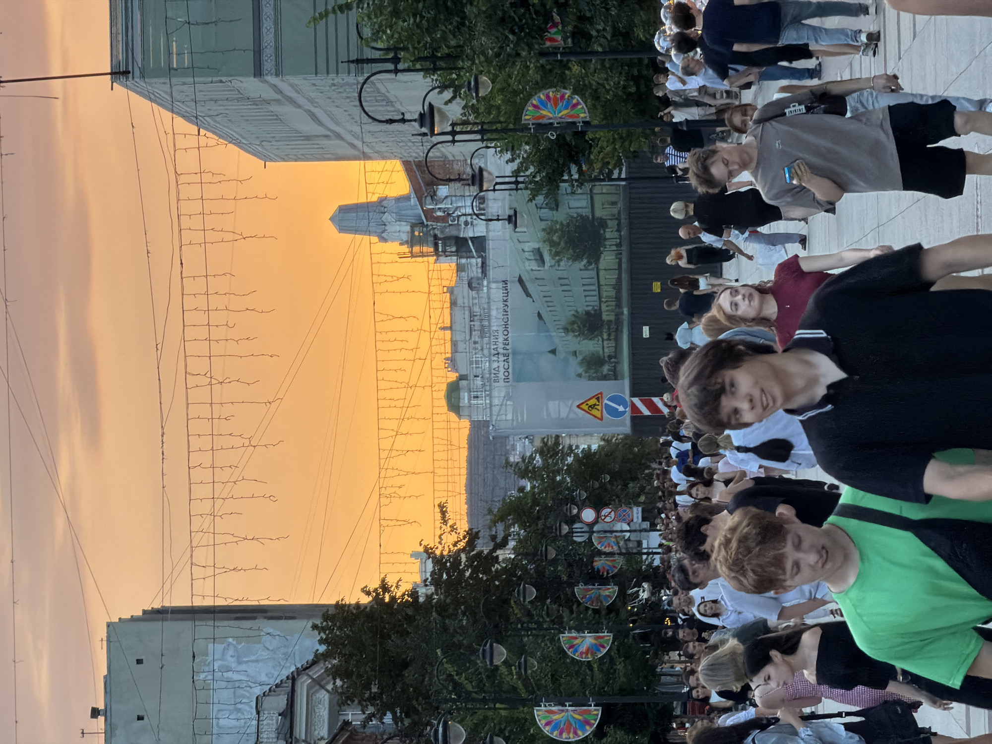 The image depicts a street scene in Saratov, Russia, during what appears to be twilight. A wide pedestrian street is filled with a crowd of people walking in both directions. The sky is a vibrant orange, characteristic of sunset or sunrise, with some clouds visible. On either side of the street, there are buildings, some of which are under construction or renovation, as indicated by scaffolding and large banners with images of buildings. Decorative string lights are hung across the street overhead. Lampposts with multiple lights line the sidewalks, and there are signs and traffic indicators along the way. Several individuals are the primary focus, including a young man in a green t-shirt in the foreground, another young man in a black polo shirt to his right, and a young man in a grey t-shirt and black shorts looking at his phone. Many other people are visible in the background, engaged in various activities such as walking, talking, and browsing on their phones. The overall atmosphere is bustling and lively, suggesting a public gathering or a popular thoroughfare.