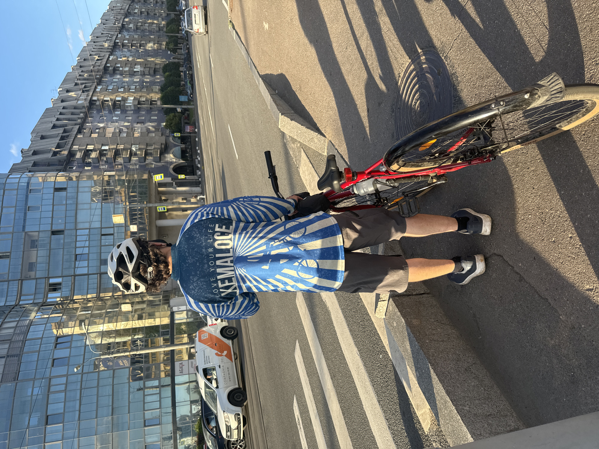 A person wearing a helmet, a blue and white long-sleeved shirt with the text "LOVE YOUR LIFE XEMALOCE" and bicycle graphics, and dark shorts stands next to a red bicycle on a sidewalk in front of a pedestrian crossing. The person is facing away from the camera, looking towards a street with cars and a modern building with a glass facade on the left and a long, blue apartment building with angled roofs on the right. The scene appears to be in Saint Petersburg, Russia, likely on a sunny day, given the clear sky and shadows. The background includes street signs, traffic, and architectural elements typical of an urban environment. The building on the left has a sign for "AGATA".