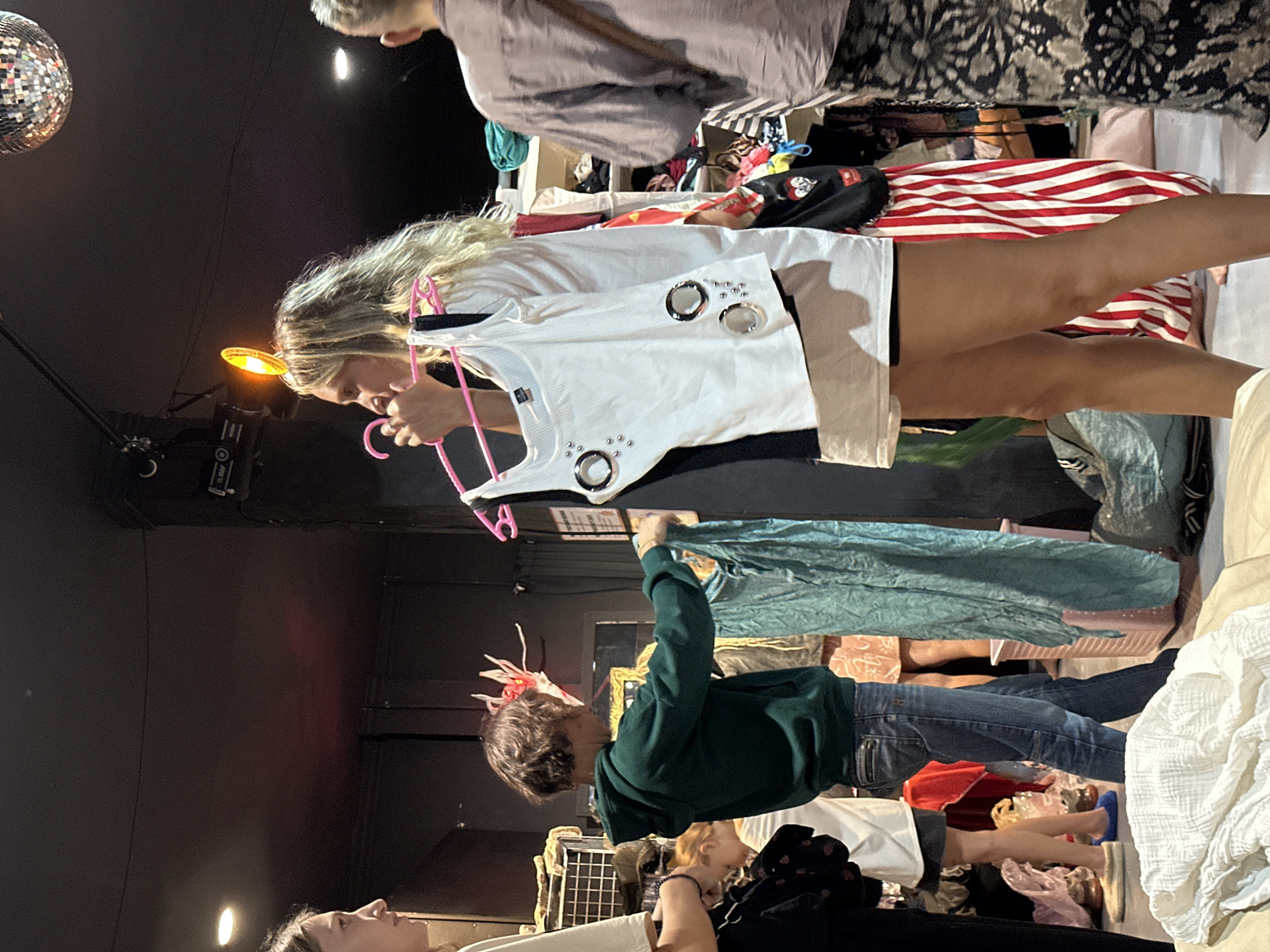 The image captures an indoor scene, likely a market or pop-up shop, filled with people browsing clothes. The setting appears dimly lit with a dark ceiling and walls, featuring a silver disco ball hanging in the upper left corner and a bright yellow-orange stage light illuminating a central area. These elements suggest an event or nighttime activity.
In the center-right, a young woman with long blonde hair, wearing a white top and a neutral-toned skirt, intently examines a white ribbed top adorned with prominent silver rings and studs, holding it up on a pink hanger. Below her, towards the lower center, another young person in a dark green hoodie and jeans holds up a piece of teal fabric, also appearing to inspect it. Various other clothing items are visible throughout the space, some neatly stacked, others piled on tables or racks, including a prominent red and white striped fabric and dark patterned textiles in the background.
Several other individuals are present; one person wearing a light brown top and patterned bottoms is partly visible behind the blonde woman, reaching into a clothes display. Another person's head is visible in the bottom left corner. The overall atmosphere suggests a casual shopping experience at a thrift store, flea market, or a fashion event. No clear text is visible on any items or signs. The activity taking place is clearly shopping for clothing, set in what appears to be an event space, consistent with the location context of Ko Pha Ngan, Thailand.