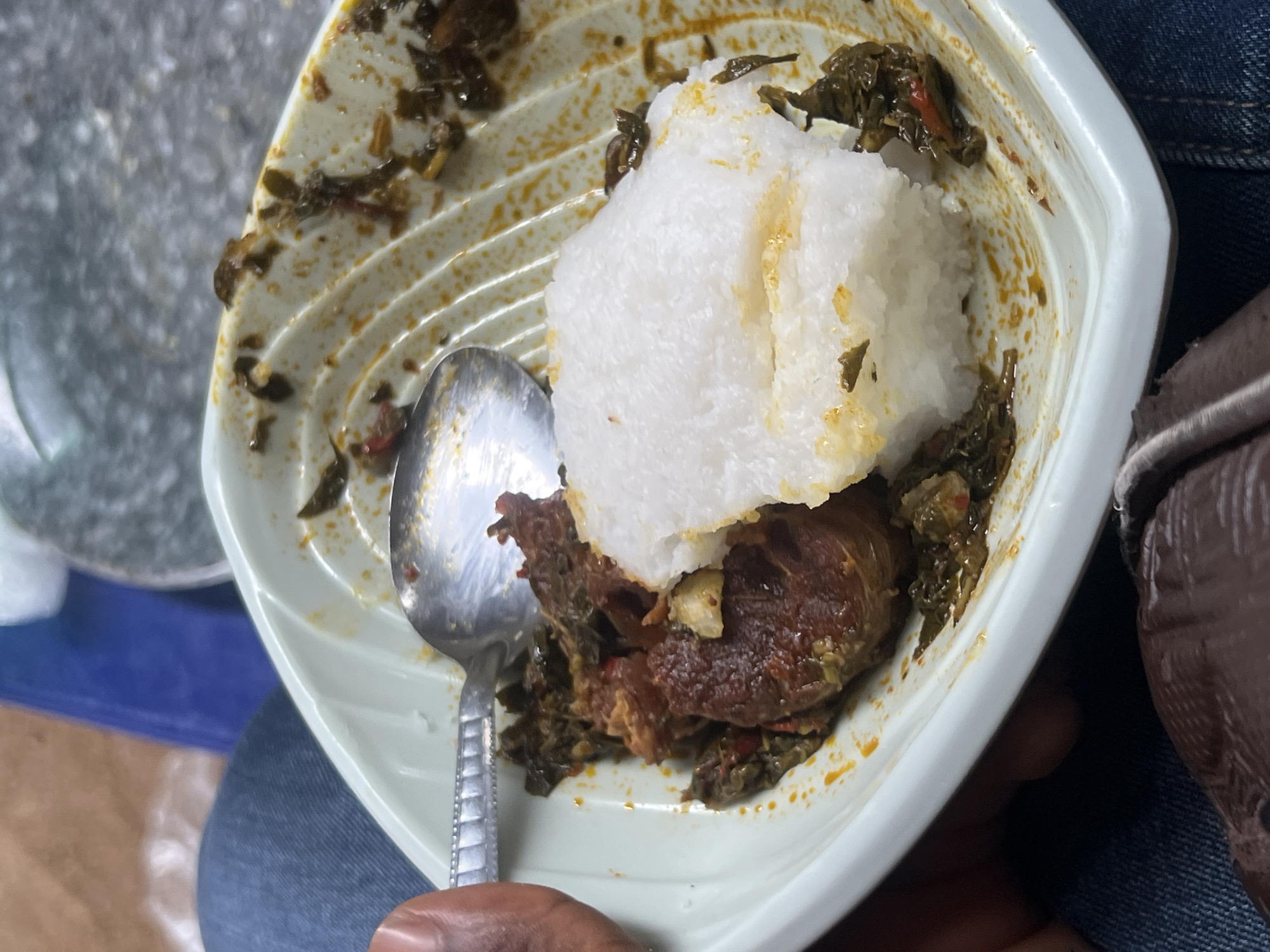 The image presents a close-up, overhead view of a meal contained within a light-colored, possibly plastic, square bowl with ridged patterns on its interior. The main subjects are the food items: a prominent white, starchy mound, characteristic of fufu, eba, or pounded yam, positioned centrally. Beneath and surrounding this staple is a dark green and brown stew, which appears to contain leafy vegetables, pieces of dark meat (likely beef or goat), and some reddish specks that could be chili or pepper. An oily, yellowish-orange residue from the stew stains the bowl's interior.
A silver metal spoon with a textured handle is placed within the bowl, indicating a meal in progress or about to be eaten. A person's hand is visible in the lower left corner, holding the spoon and the bowl. The person is wearing blue denim jeans. On the right side of the frame, a blurred section of brown fabric or a bag is visible. The background is out of focus, showing a grey, textured surface and a hint of blue, suggesting an informal setting where the meal might be held on a lap. The cuisine is consistent with West African dishes, aligning with the location context of Jalingo, Nigeria. There is no visible text in the image.