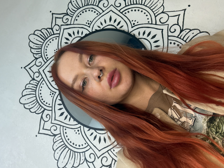 A woman with long, vibrant red hair is the focal point, angled slightly towards the camera. She has fair skin with freckles, green eyes, and piercings in her nose. Her expression is neutral. Behind her is a white wall featuring a large, black mandala design. A round, grey object is partially visible behind her head, framed by the mandala. She wears a brown shirt with a visible pattern and a light-colored jacket or sweater.