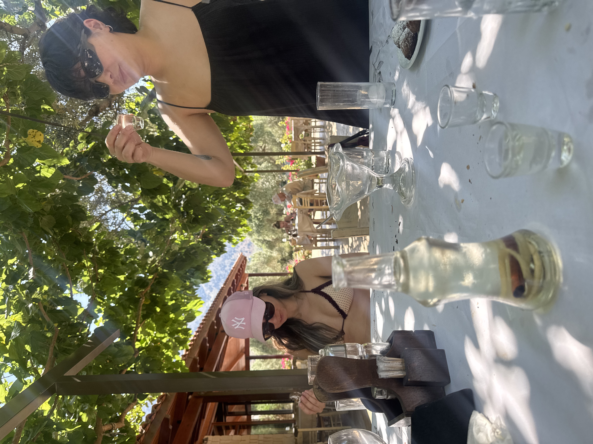 The image depicts an outdoor dining scene in Dramia, Greece, under the canopy of a large tree providing dappled shade. Two women are seated at a white-clothed table in the foreground.
On the right, a woman with short dark hair and dark sunglasses wears a black spaghetti-strap top. She holds a small, clear shot glass containing a clear liquid at eye level, appearing to inspect or prepare to drink from it. A tattoo is visible on her upper left arm.
On the left, a woman with long, light brown hair wears a pink baseball cap with a white "NY" logo and dark sunglasses. She is dressed in a light-colored, possibly beige, polka-dot bikini top with dark trim. Her right hand is raised, holding a small, indistinct object.
The white tablecloth is cluttered with various clear glass items, including an ornate stemmed dessert cup, taller drinking glasses, and two empty shot glasses in the lower right. A clear glass bottle containing a cinnamon stick and a small piece of light-colored material sits prominently in the foreground. A wooden caddy holding condiments and napkins is positioned between the two women. On the right side of the table, a small white plate holds several dark, powdered cookies.
The background features additional tables with patrons, partially obscured by olive trees and flowering bushes. A building with a tiled roof and wooden structural elements is visible to the left. Distant mountains rise under a clear sky. Strong sunlight filters through the tree canopy, casting bright patches and distinct shadows across the table and subjects.