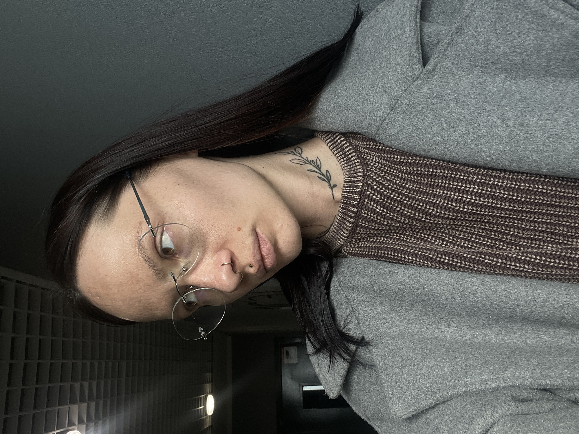 A person is indoors, photographed from a low angle. The individual has dark hair, fair skin, and wears round eyeglasses. A nose piercing is visible. A small tattoo of a plant is on the neck. They are wearing a brown knit sweater under a grey coat. The background shows a grid-like pattern on the left, possibly a wall, and a darker area suggesting a hallway or doorway straight ahead. No specific details indicate this is Izhevsk, Russia.