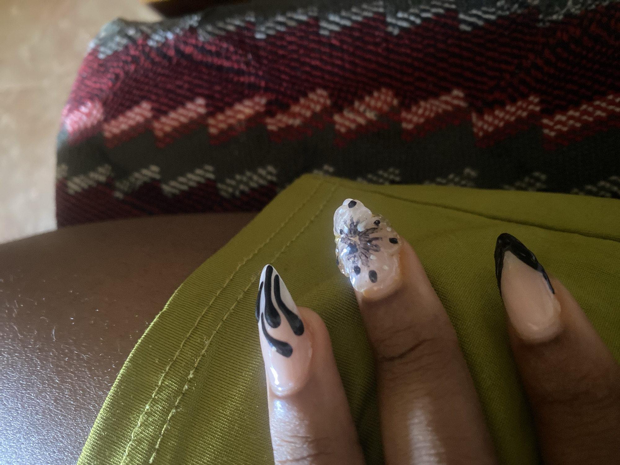 This media file features a close-up view of a person's hands with intricately designed fingernails. The nails have a light, natural base color with black and white designs. One nail displays a flame-like pattern in black on a nude base. Another nail has a design resembling a flower or butterfly with black dots and delicate black lines on a white background, adorned with a clear gel or resin. The third visible nail has a classic French tip design with black tips. The fingers are resting on a textured, olive-green fabric with visible stitching along the edge. In the background, a portion of a patterned textile is visible, featuring a geometric design with shades of deep red, dark green, and white. The overall lighting suggests an indoor setting, possibly with natural light. The scene focuses on the detailed nail art, highlighting the artistry and personal expression. No text is visible in the image. The context suggests this might be a display of personal grooming or artistic expression in Jos, Nigeria.