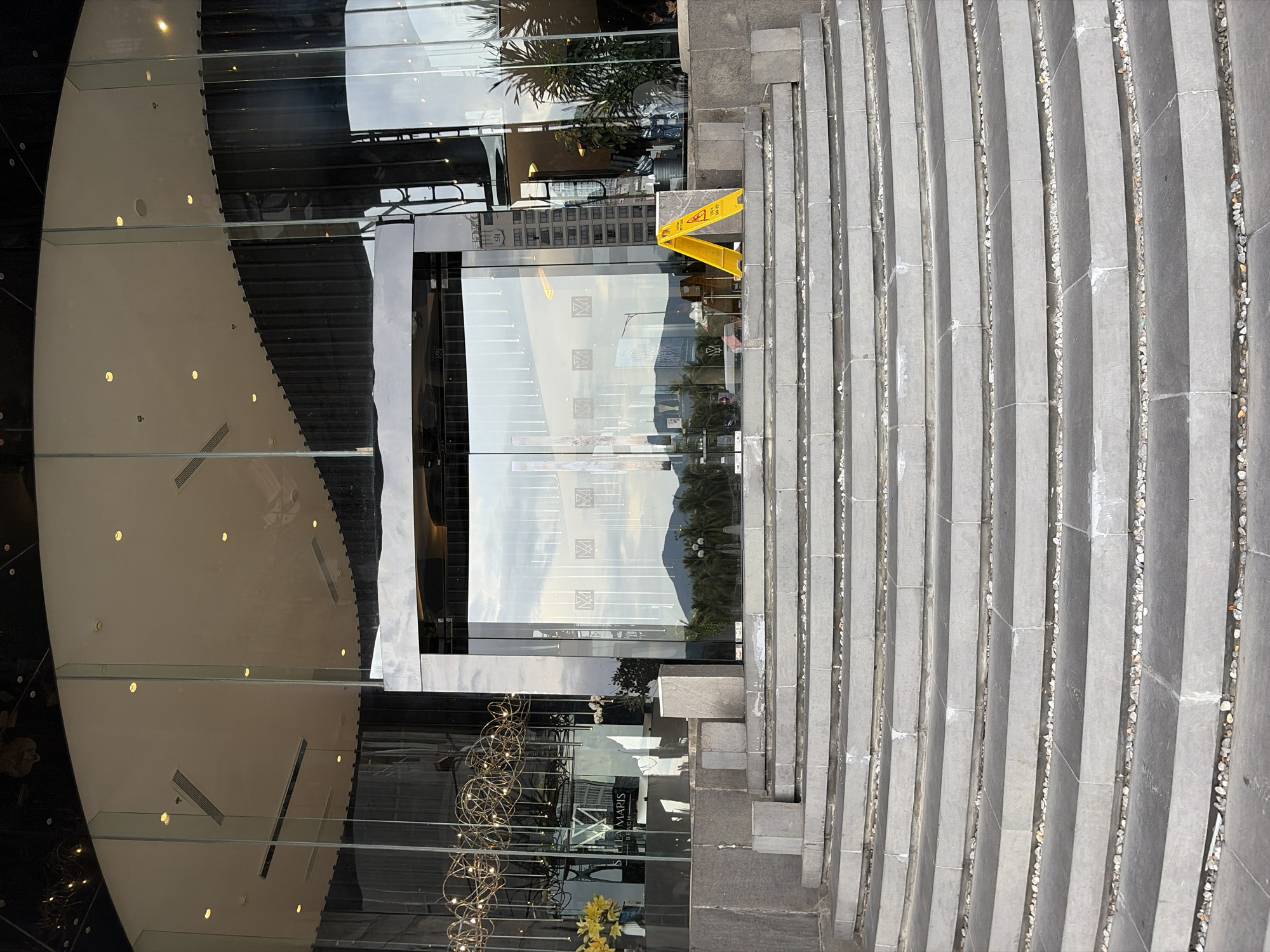 The image shows the entrance to the Stella Maris Beach Hotel in Da Nang, Vietnam. A wide, curved set of stone stairs leads up to a modern glass entrance. The doors are flanked by large glass panels, and the hotel's name is visible on a sign inside. Outside the entrance, a yellow "wet floor" sign is propped up. The sky is visible through the glass doors, reflecting palm trees and a distant mountain range.