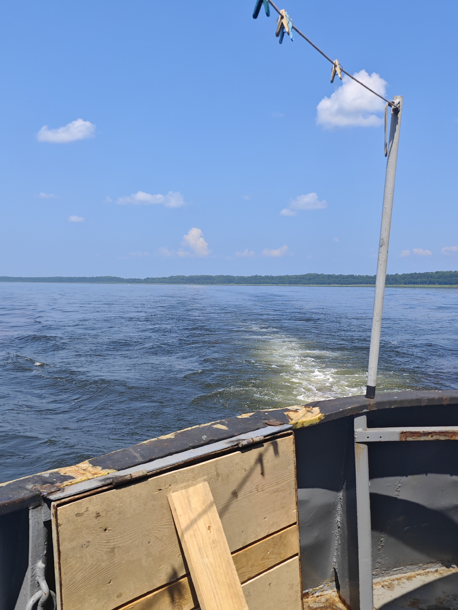 The image depicts a view from the back of a boat moving through a body of water under a clear blue sky with scattered white clouds. The water shows a visible wake and churned foam behind the boat. The foreground shows the weathered deck of the boat with some wooden planks. A clothesline with several clothespins is strung across the upper right portion of the image. In the distance, a treeline of dense green foliage borders the horizon. The scene appears to be set on a calm, sunny day, likely in a rural or natural setting. The specific location in the city of Погодаево, Russia, is not discernible from the image itself, as there are no identifiable landmarks or structures that would indicate this specific geographic area.