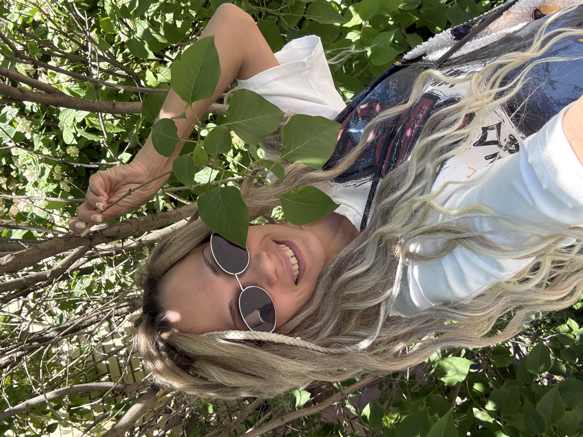 In Rusanovo, Russia, a female individual with long, wavy blonde hair, partially styled in braids, is captured in an outdoor setting under bright sunlight. She is wearing sunglasses with oval metallic frames and a white crew-neck t-shirt, over which a patterned garment or accessory with dark, white, and red/pink abstract designs is visible. Her right hand, featuring light-colored nail polish, firmly grips a thin tree branch adorned with green leaves, positioned in front of her face. The individual displays a wide smile, revealing her teeth, which exhibit a yellowish discoloration. The background consists of dense green foliage, indicative of a bush or small tree, interspersed with thin, visible branches. Patches of small white flowers are present within the background vegetation. A light-colored, possibly brick, wall is partially visible in the upper left portion of the frame. Strong shadows are cast, consistent with a sunny outdoor environment.