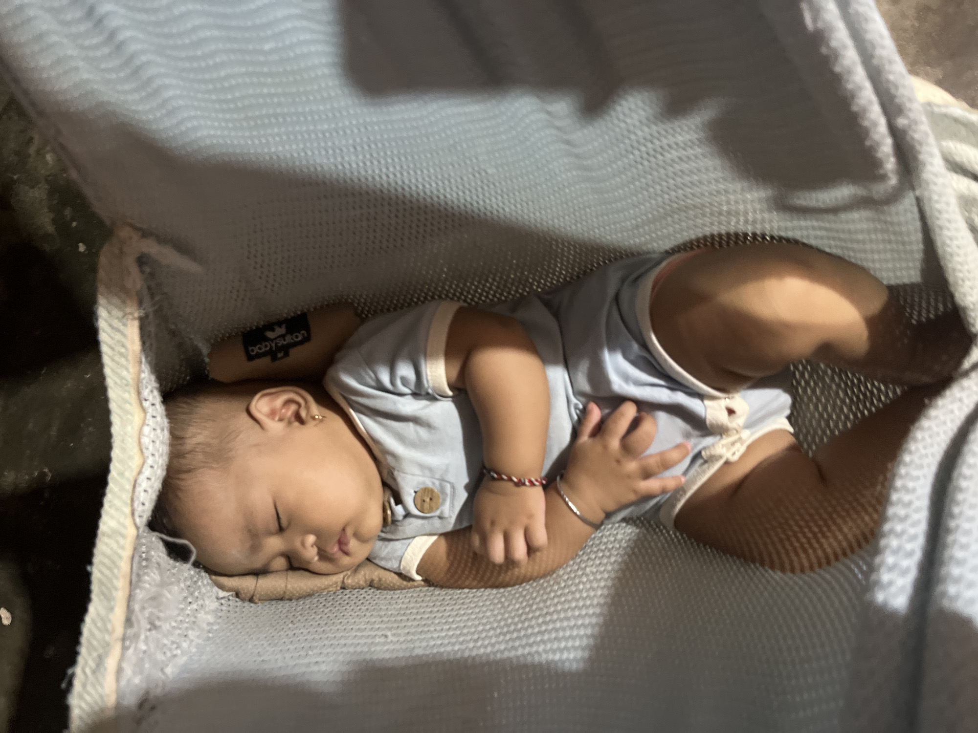 A young child is asleep in a light blue hammock. The child is wearing a light blue onesie with a wooden button closure on the front. The child has a small gold earring in their right ear and two bracelets on their left wrist, one with red and white beads and the other a thin silver band. The hammock is made of a mesh-like fabric and appears to be suspended in a dark, indoor environment. The background is out of focus but suggests a natural or utilitarian surface. The scene is illuminated by a soft light source, casting shadows across the hammock and the child. The image was captured at an undisclosed location within Kruengmate, Indonesia.