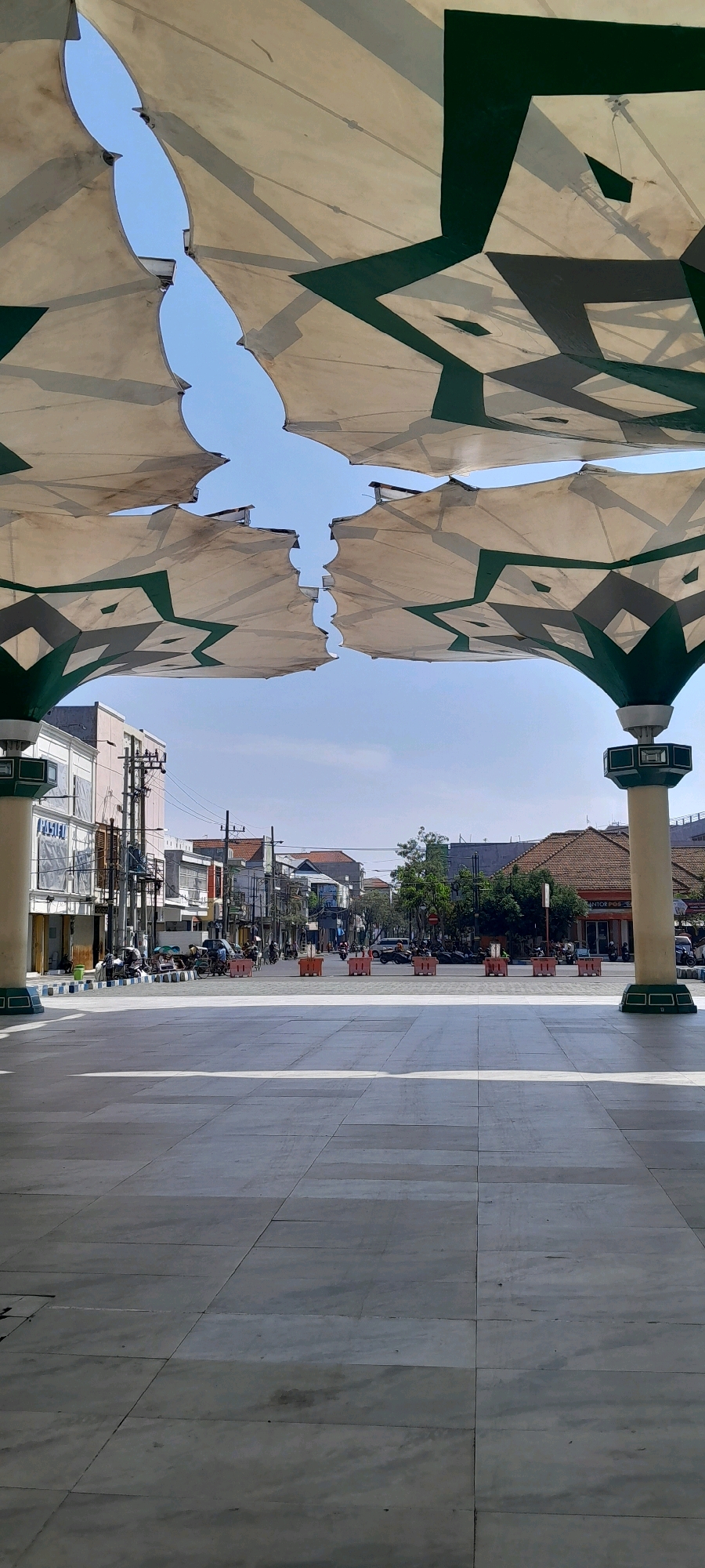 The image captures an outdoor scene, likely a public square or plaza, in a city. In the foreground, a large, tiled surface dominates, reflecting the bright sunlight. Overhead, a large, white fabric canopy with green geometric patterns provides shade, creating a segmented view of the sky. The canopy is supported by large, pillar-like structures.
Beyond the plaza, a street is visible with buildings lining both sides. These buildings appear to be a mix of commercial establishments and residences, with signs and storefronts. The street is moderately busy with traffic, including motorcycles and cars. Several individuals are present, some on motorcycles and others standing or walking near the buildings. Orange traffic barriers are placed along the street. Trees and utility poles are also part of the urban environment. The overall impression is a sunny day in a bustling urban area, with the distinctive canopy offering a focal point.