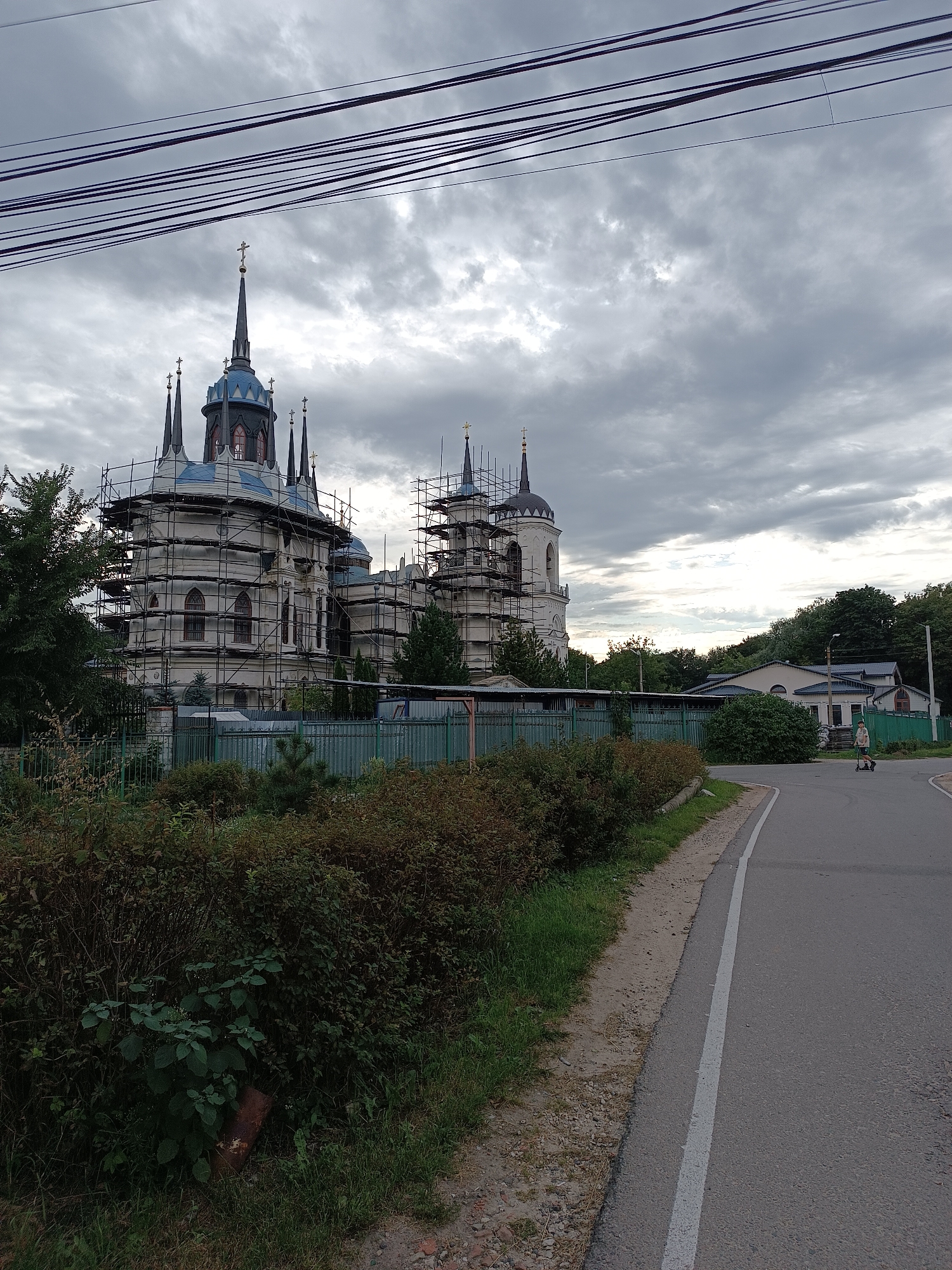 The image depicts the Church of the Ascension of the Lord in Bykovo, Russia, under scaffolding for renovation. The imposing church, with its distinctive blue domes and multiple spires topped with crosses, dominates the central and left portions of the frame. Scaffolding encases much of the building's exterior, suggesting ongoing construction or repair work.
To the right of the church, a road curves away from the viewer. A person is visible on a scooter on this road, heading away from the church. Adjacent to the road and church is a green fence and dense green foliage, with some bushes and trees. Further in the background, on the right side, are several low-rise buildings with dark roofs. The sky above is overcast with dramatic, dark clouds, contributing to a somber atmosphere. Overhead power lines are prominently stretched across the top of the image. The foreground is filled with lush green bushes and grass, with a section of dirt and gravel shoulder along the road.